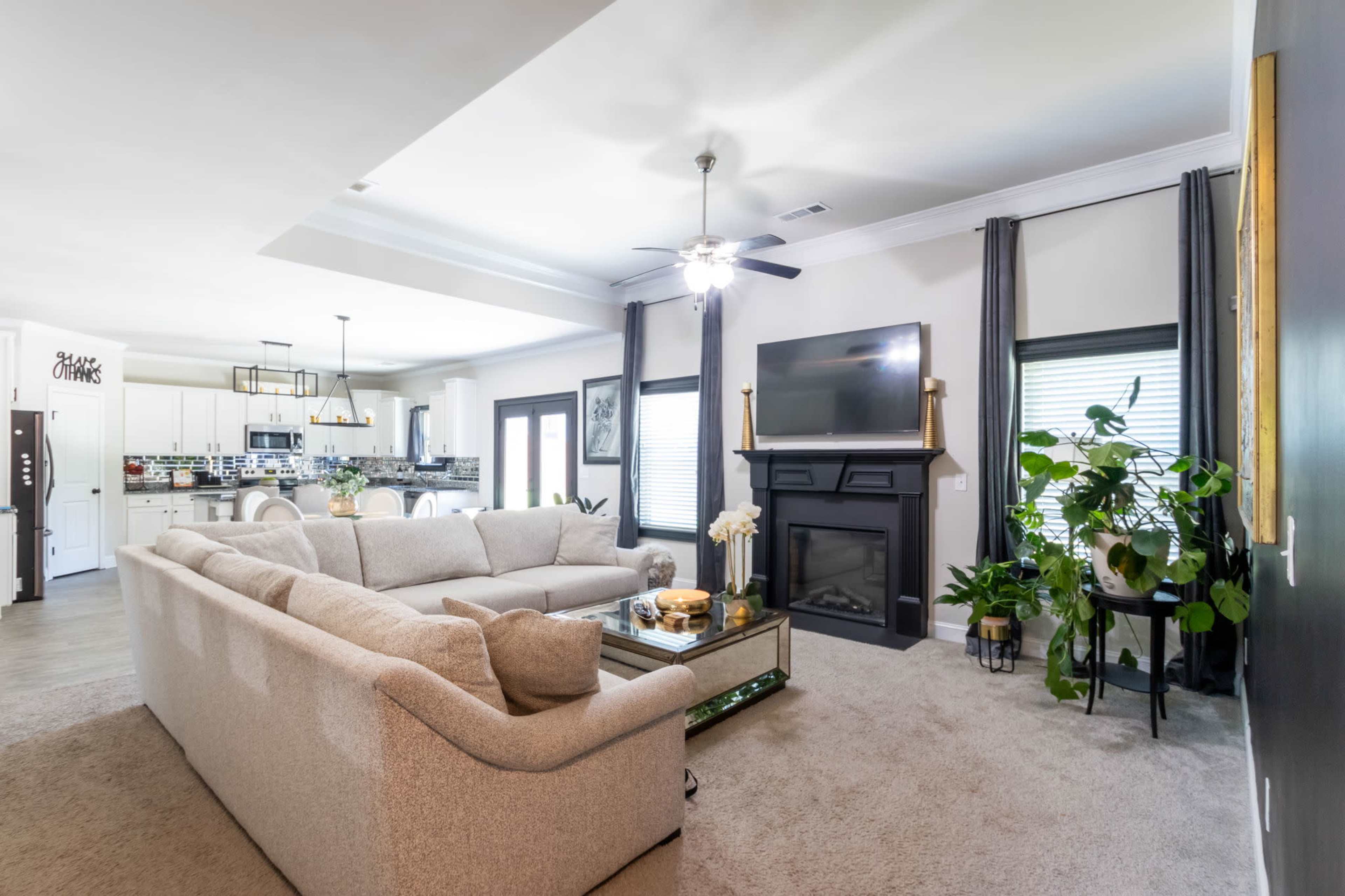 The image shows a modern living room with a large sectional sofa, a coffee table, and a black fireplace, while a kitchen area with light-colored cabinetry is visible in the background.