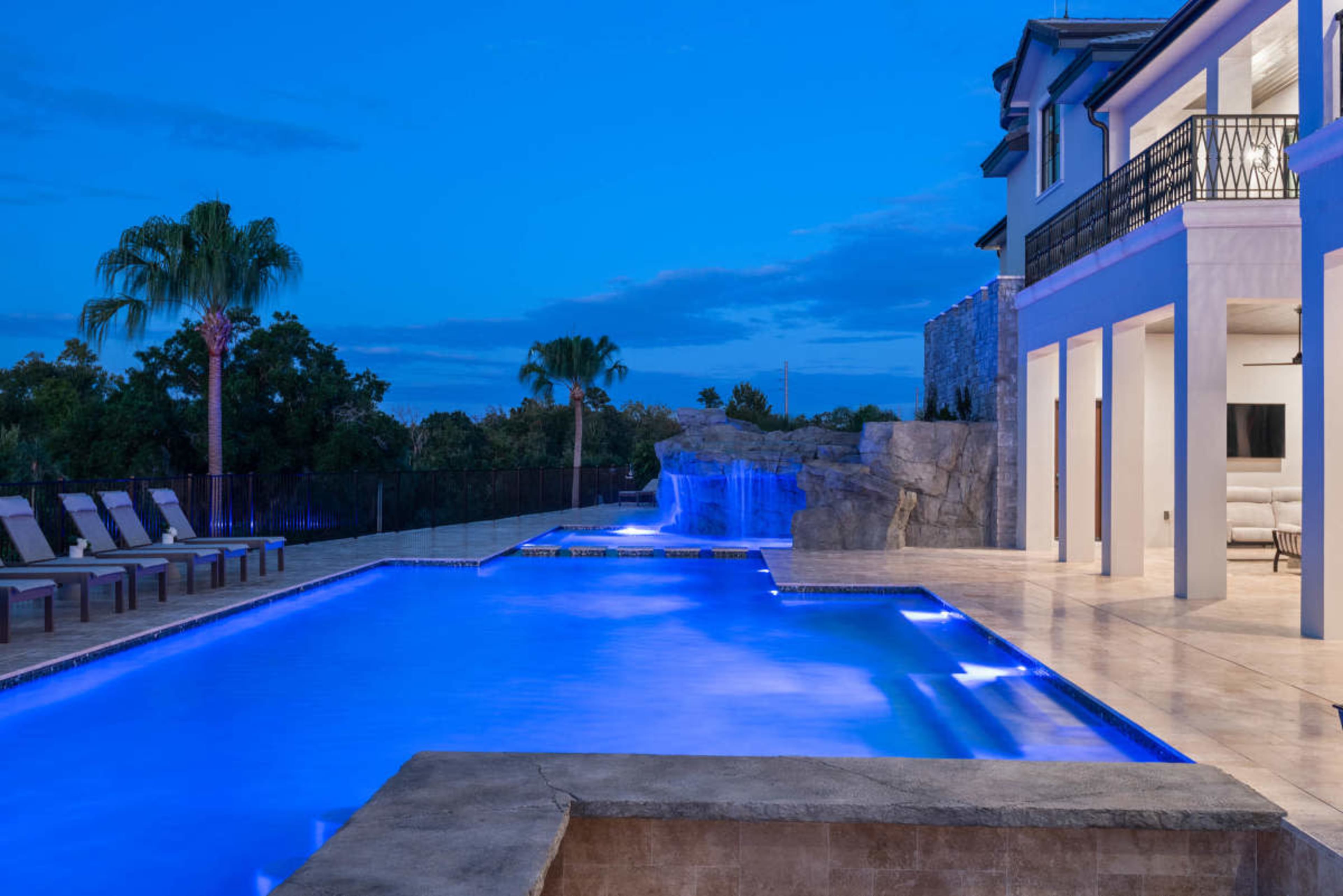 The image shows a modern outdoor pool illuminated with blue lighting alongside a waterfall feature and a two-story house at dusk.