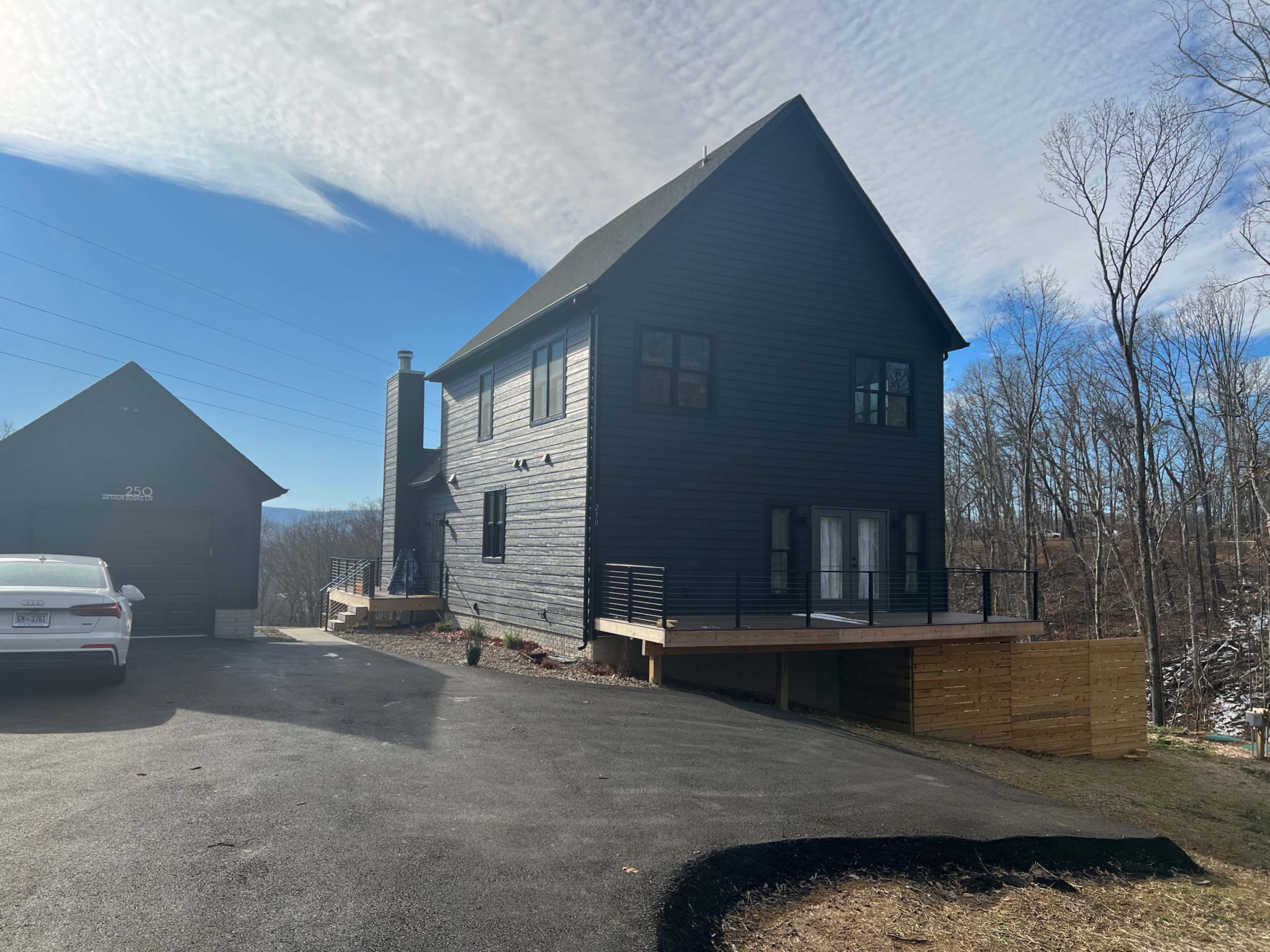 A modern black house with a wooden deck is situated on a slope, with a garage visible nearby.
