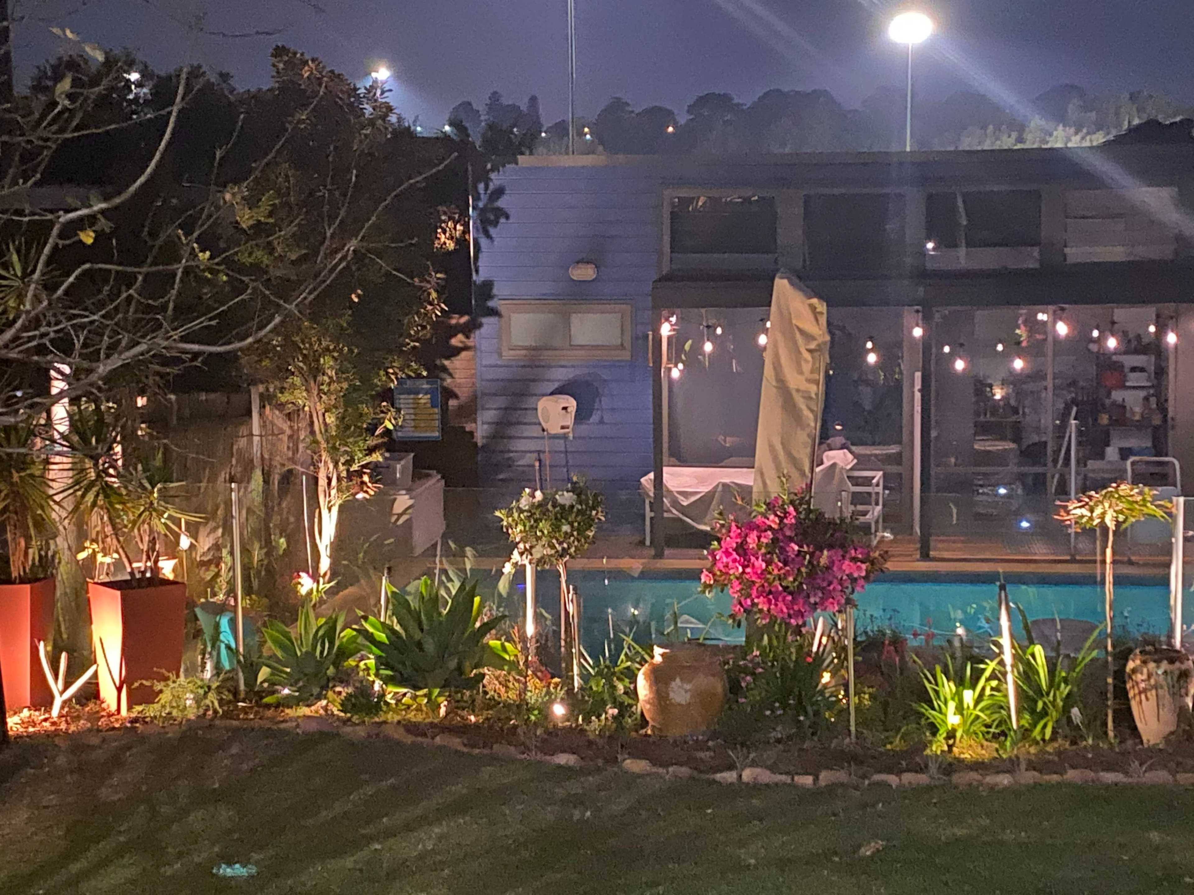 A backyard scene at night features a well-lit pool area surrounded by potted plants and a modern blue house.