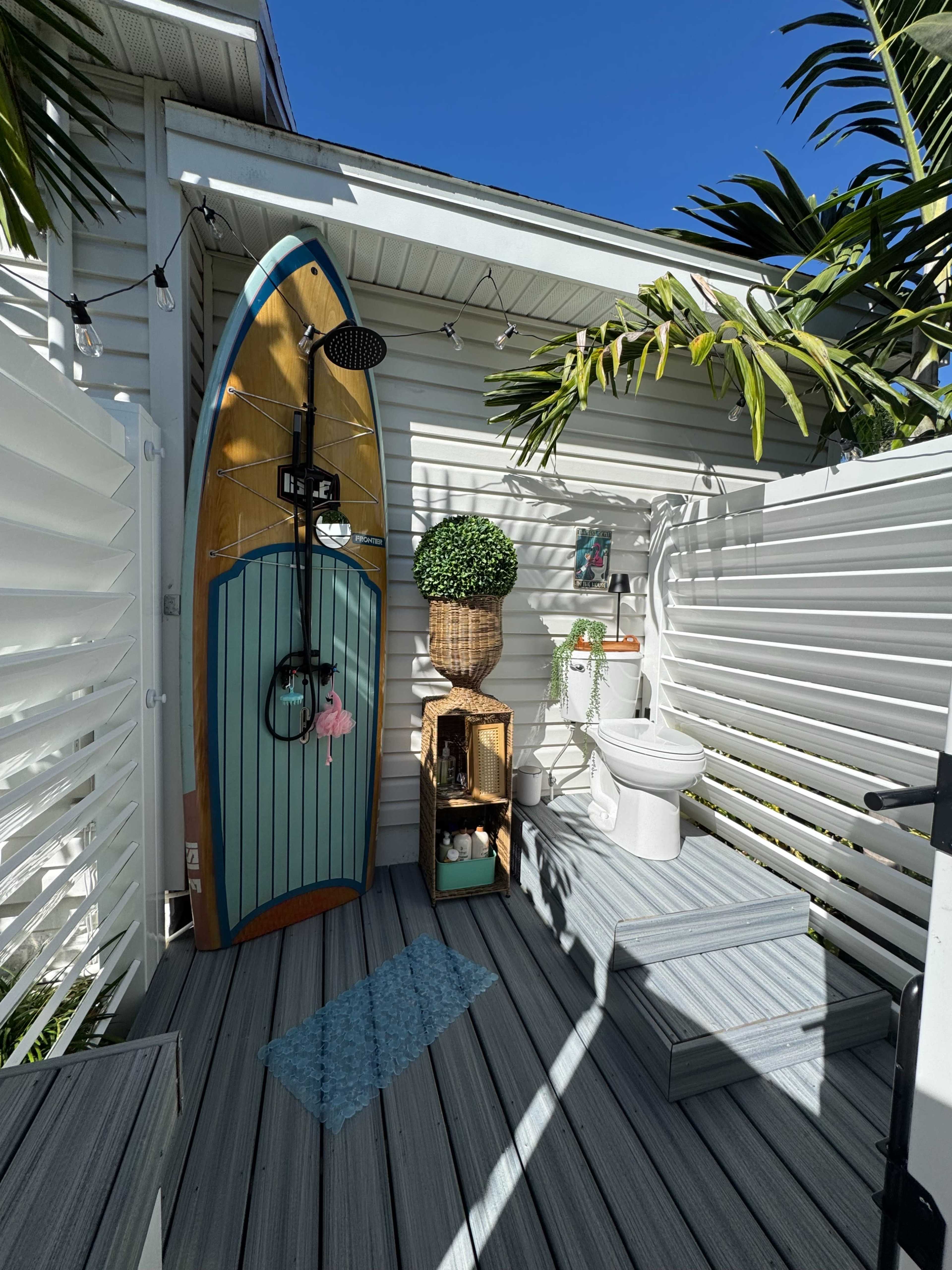 The image shows an outdoor shower area with a surfboard-shaped shower fixture, a toilet, a wooden storage unit, and tropical plants surrounded by white slatted fencing.