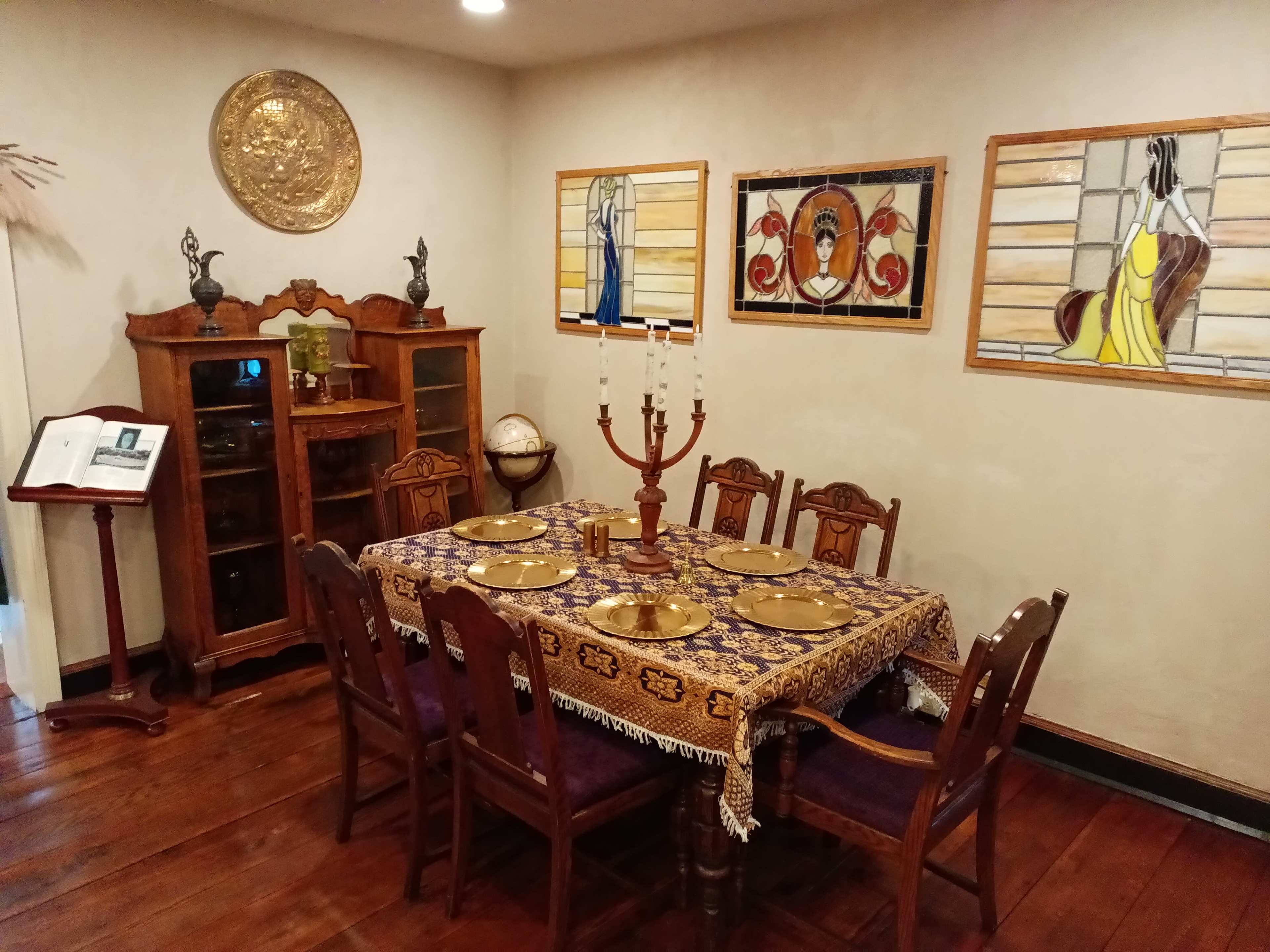 A wooden dining table is set with plates and candles, surrounded by eight chairs in a room featuring stained glass artwork and a large wooden cabinet.