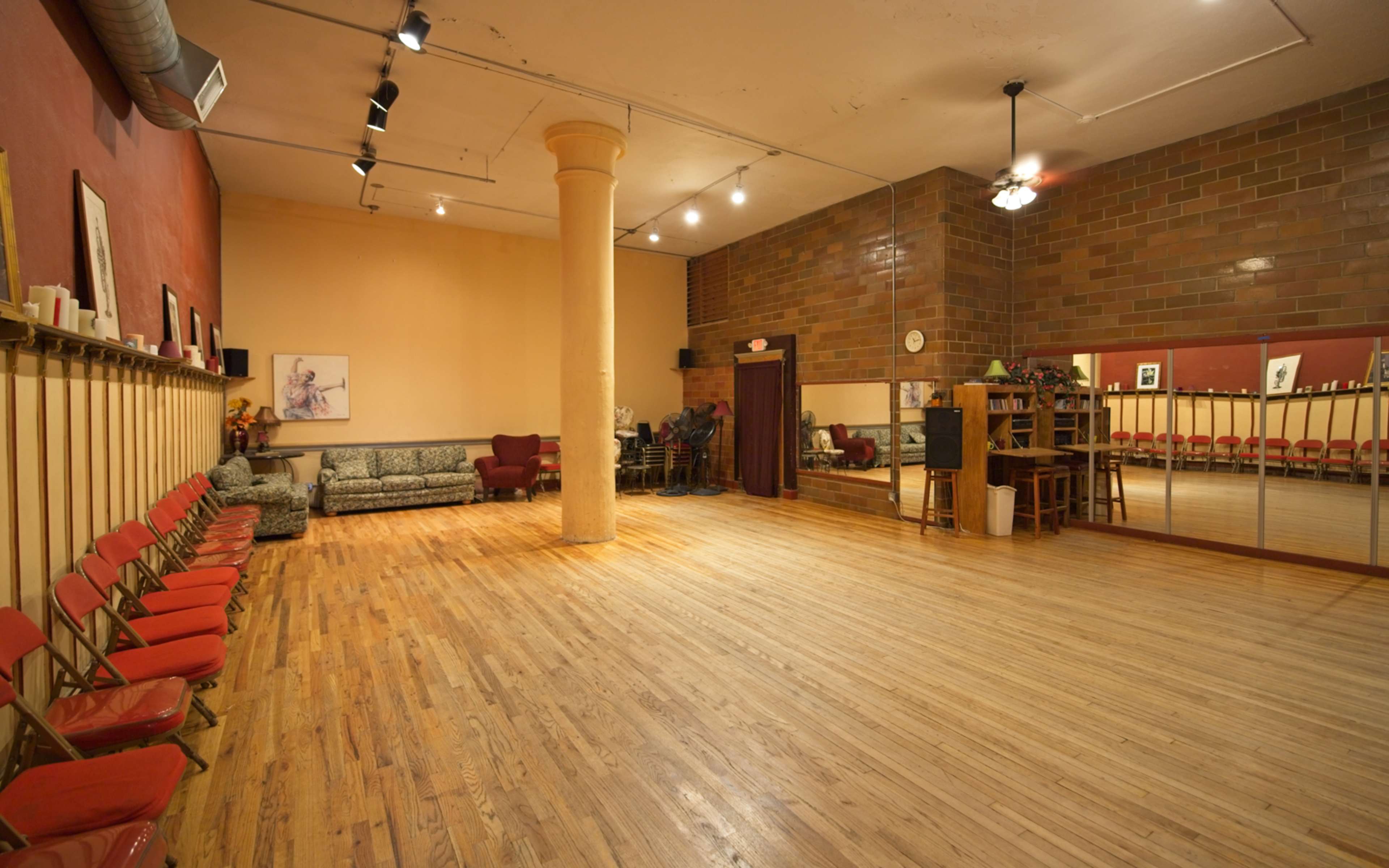 A spacious, well-lit dance studio featuring wooden floors, mirrors along one wall, and seating along the opposite side.