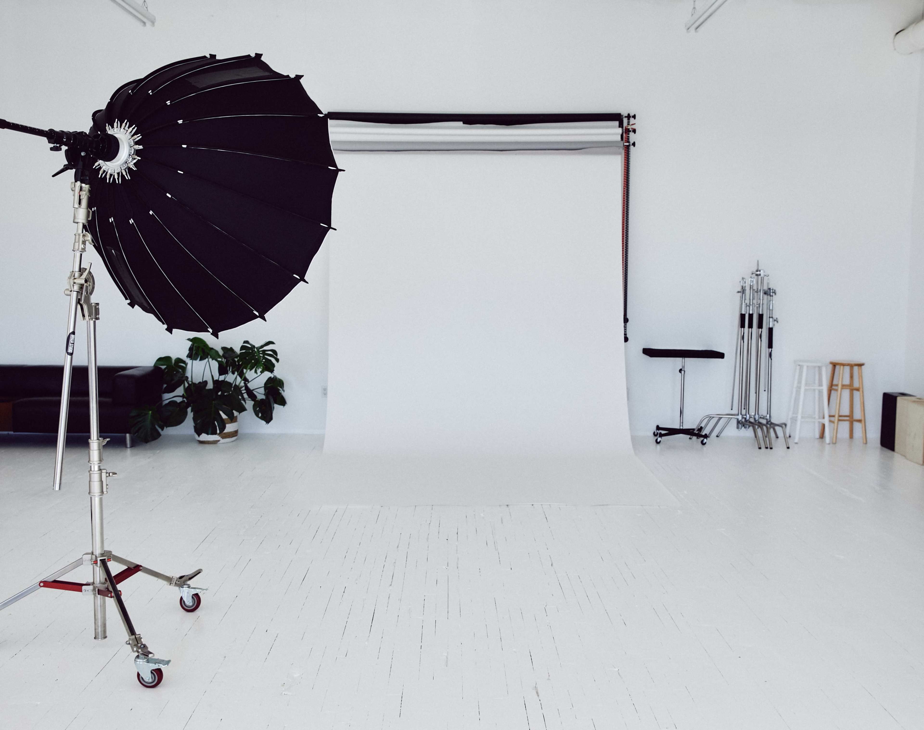 A photography studio features a large black umbrella light, a white backdrop, and various equipment on a clean, white floor.