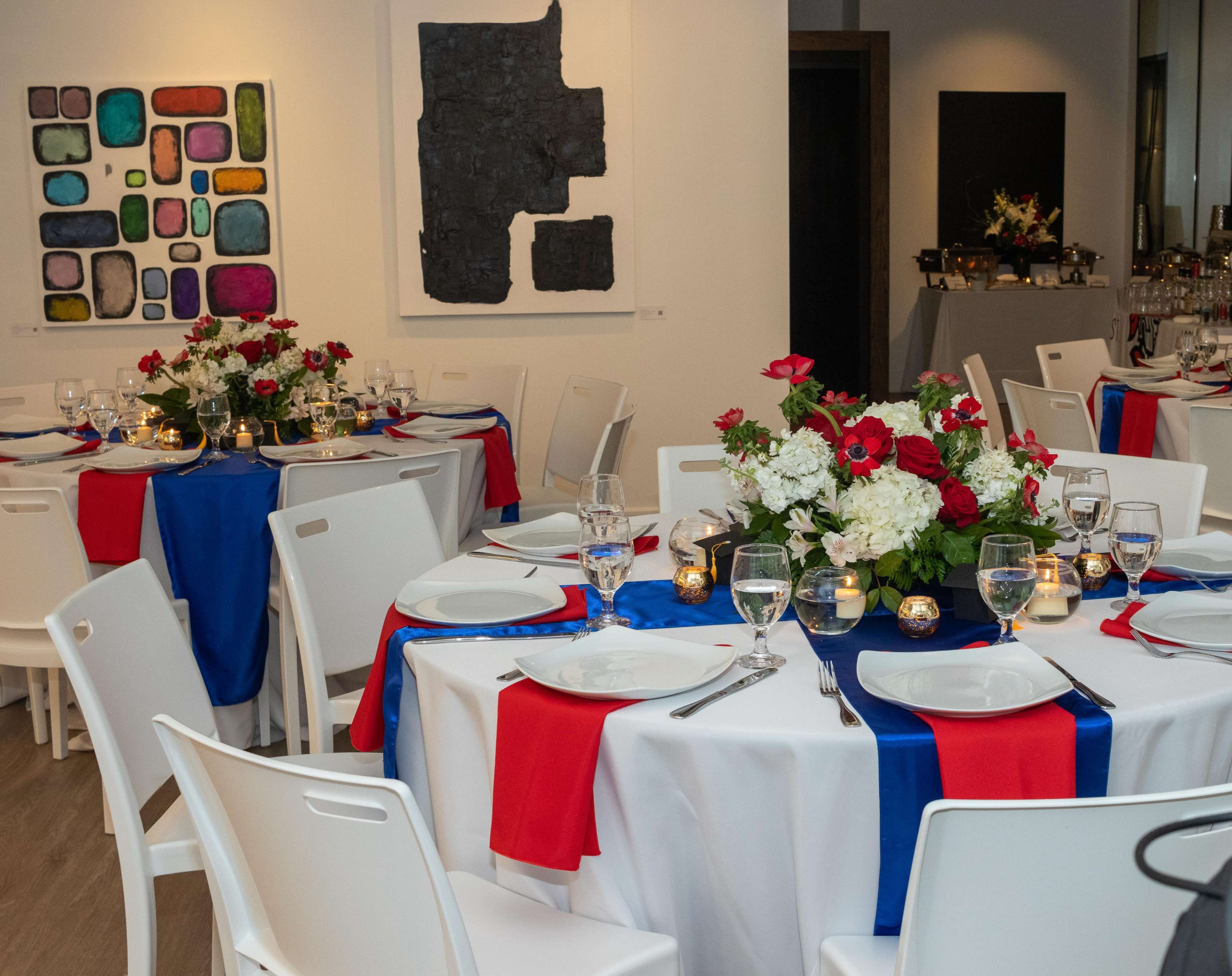 The image shows a dining setup with round tables covered in white tablecloths, adorned with blue and red fabric accents, and decorated with floral centerpieces, in a room featuring modern art on the walls.