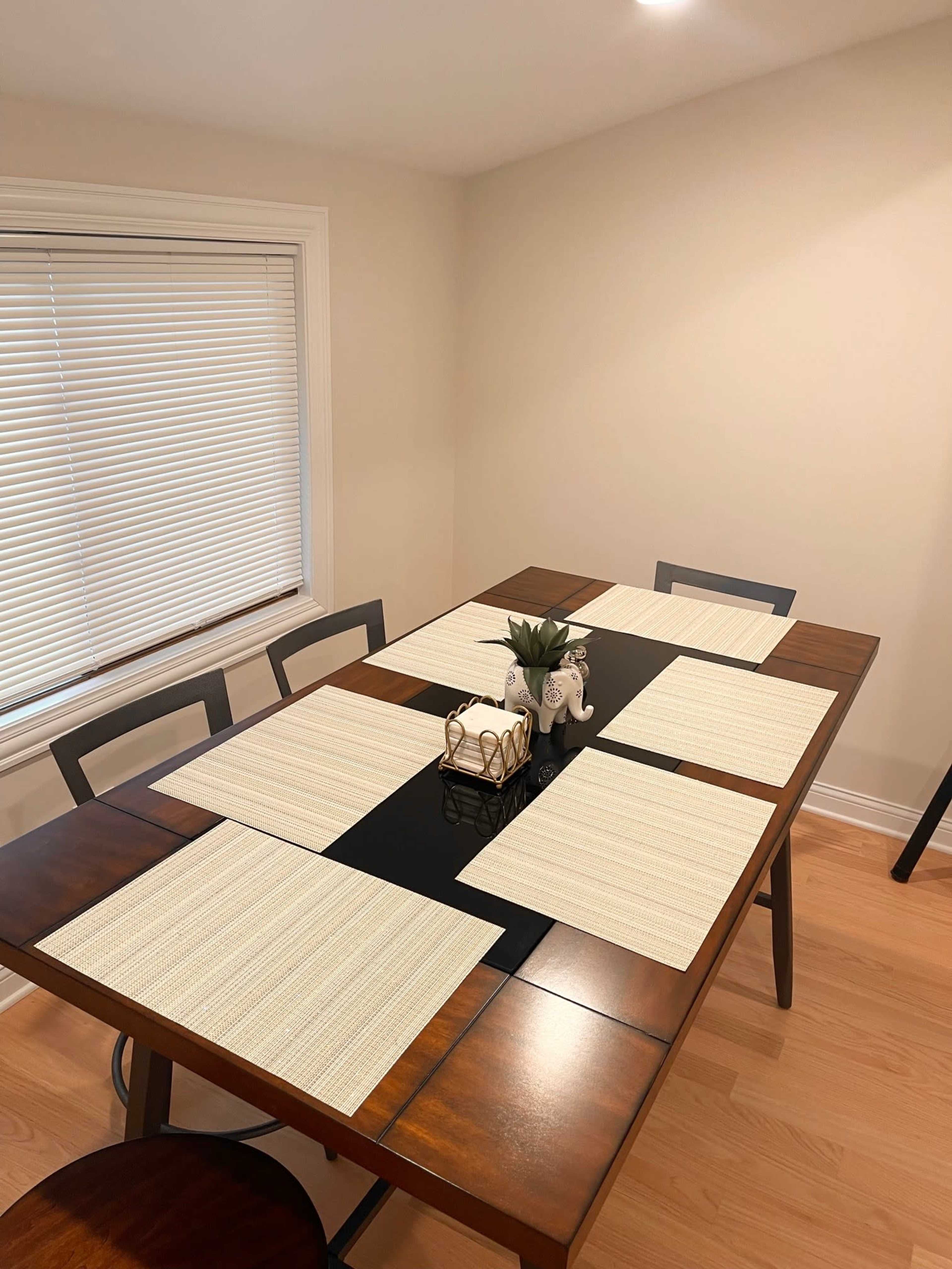A wooden dining table is set with six placemats, featuring a central decorative arrangement of a small plant and figurines.