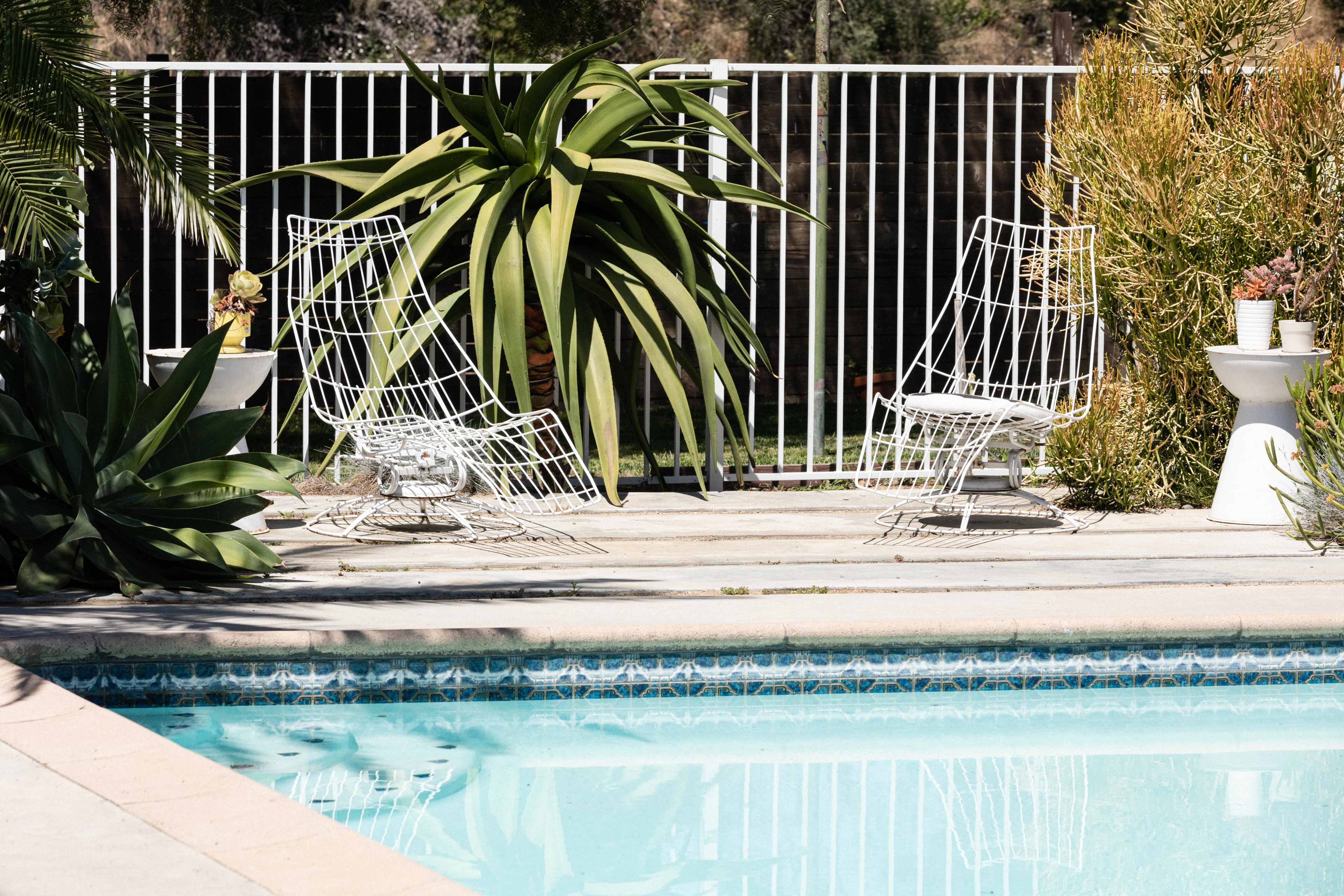 Two white wire chairs are positioned near a swimming pool surrounded by greenery and a white fence.