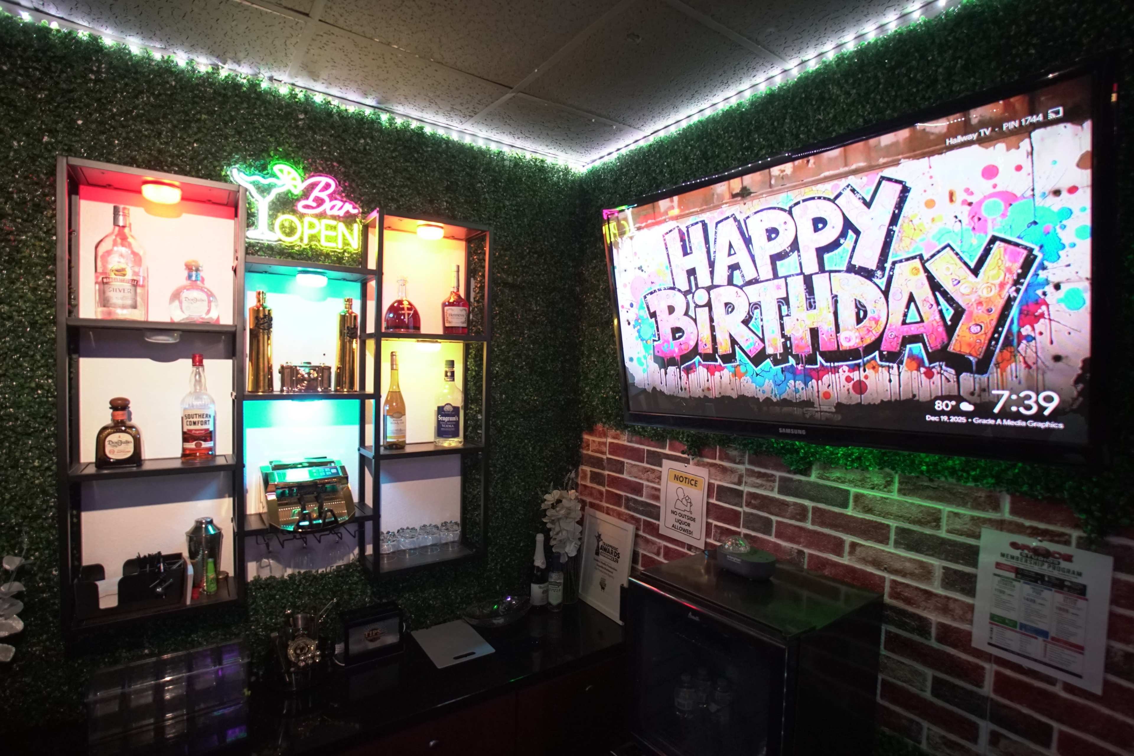 The image shows a festive bar area with a variety of liquor bottles displayed on shelves, a "Bar Open" neon sign, and a television screen displaying a "Happy Birthday" message against a colorful background.