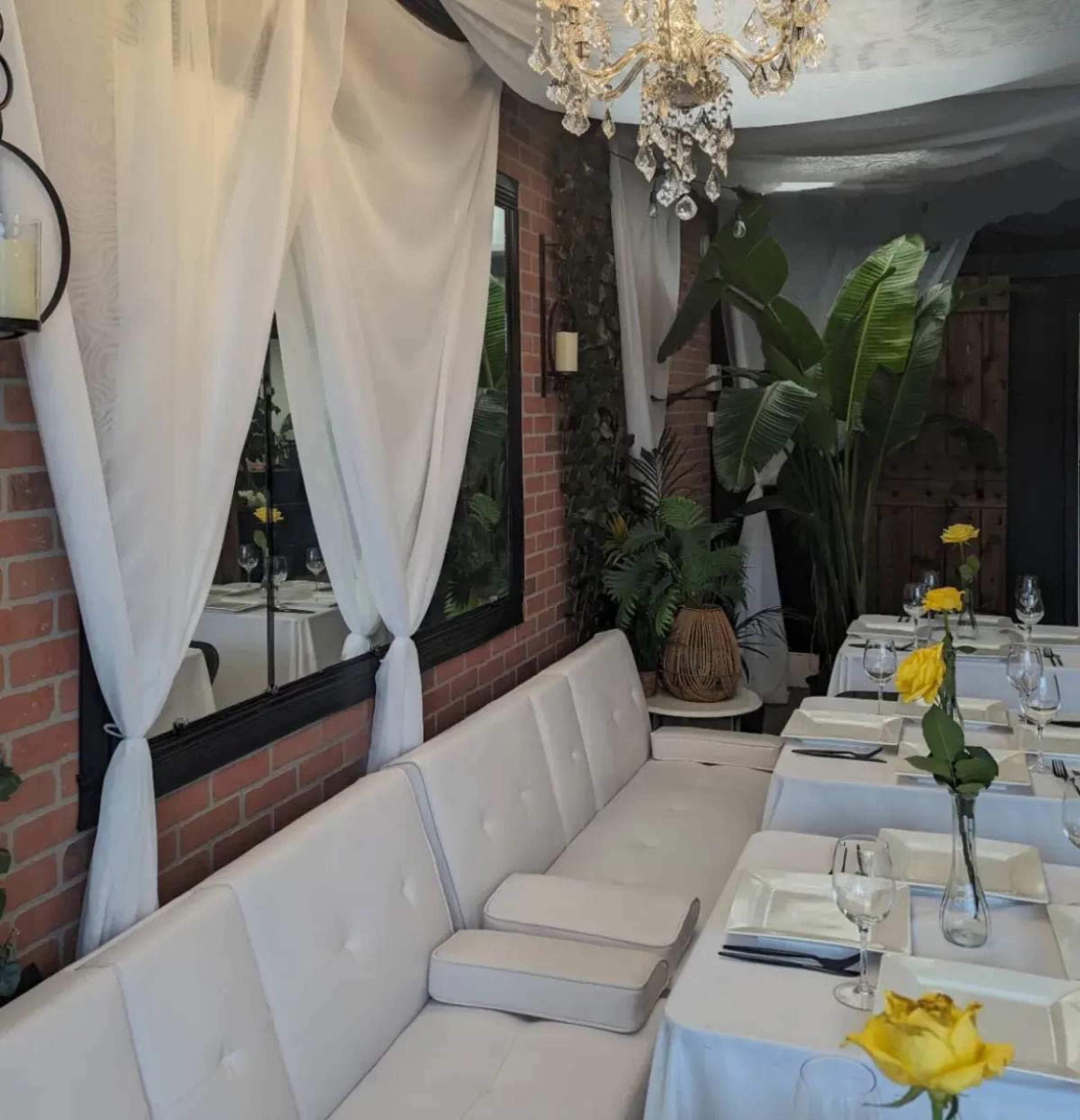 A restaurant interior featuring a white upholstered bench, tables set with plates and glasses, and plants placed throughout the space.