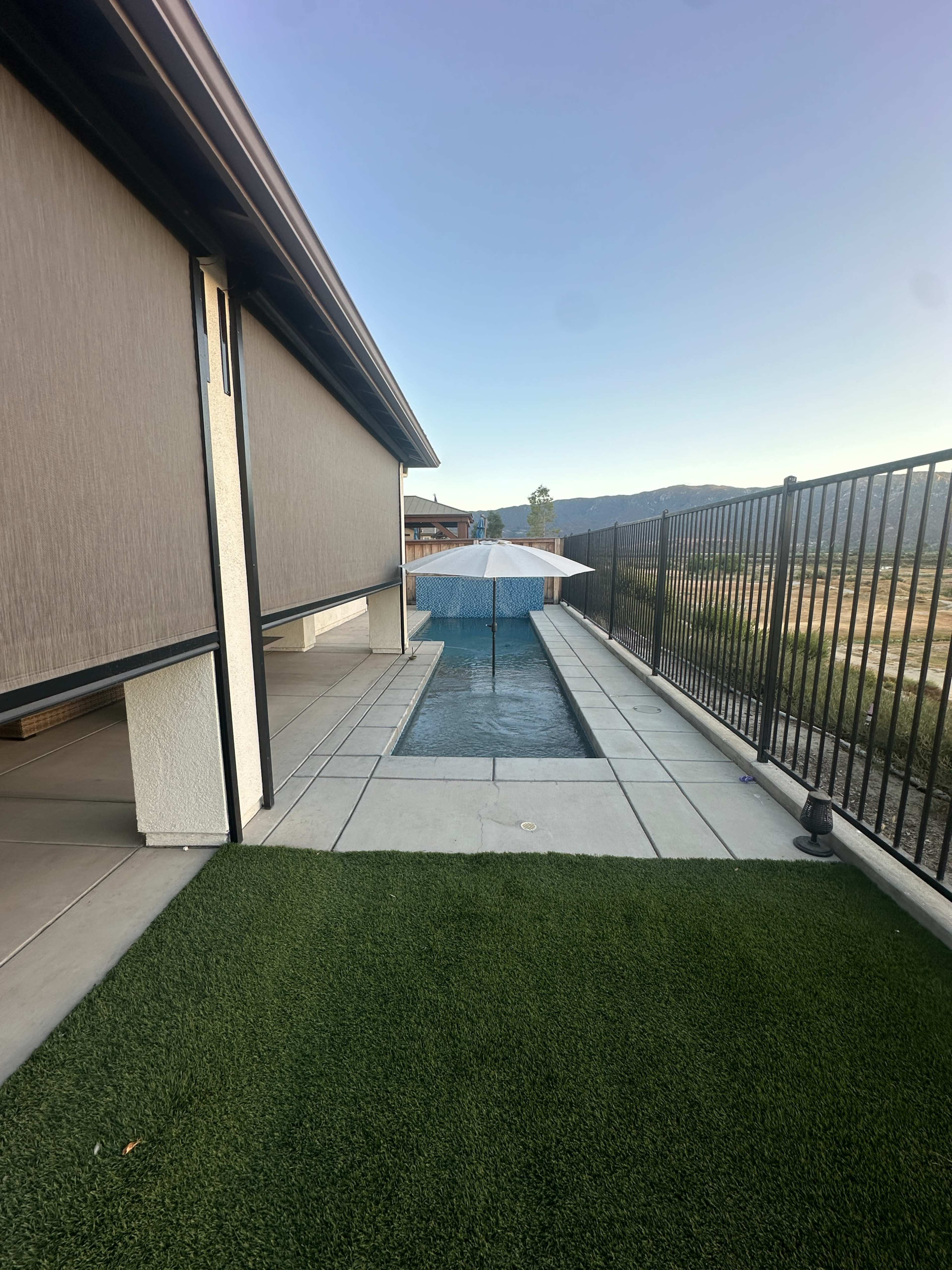 The image shows a narrow pool bordered by a concrete walkway and a grass area, with an umbrella positioned over the water and mountains visible in the background.