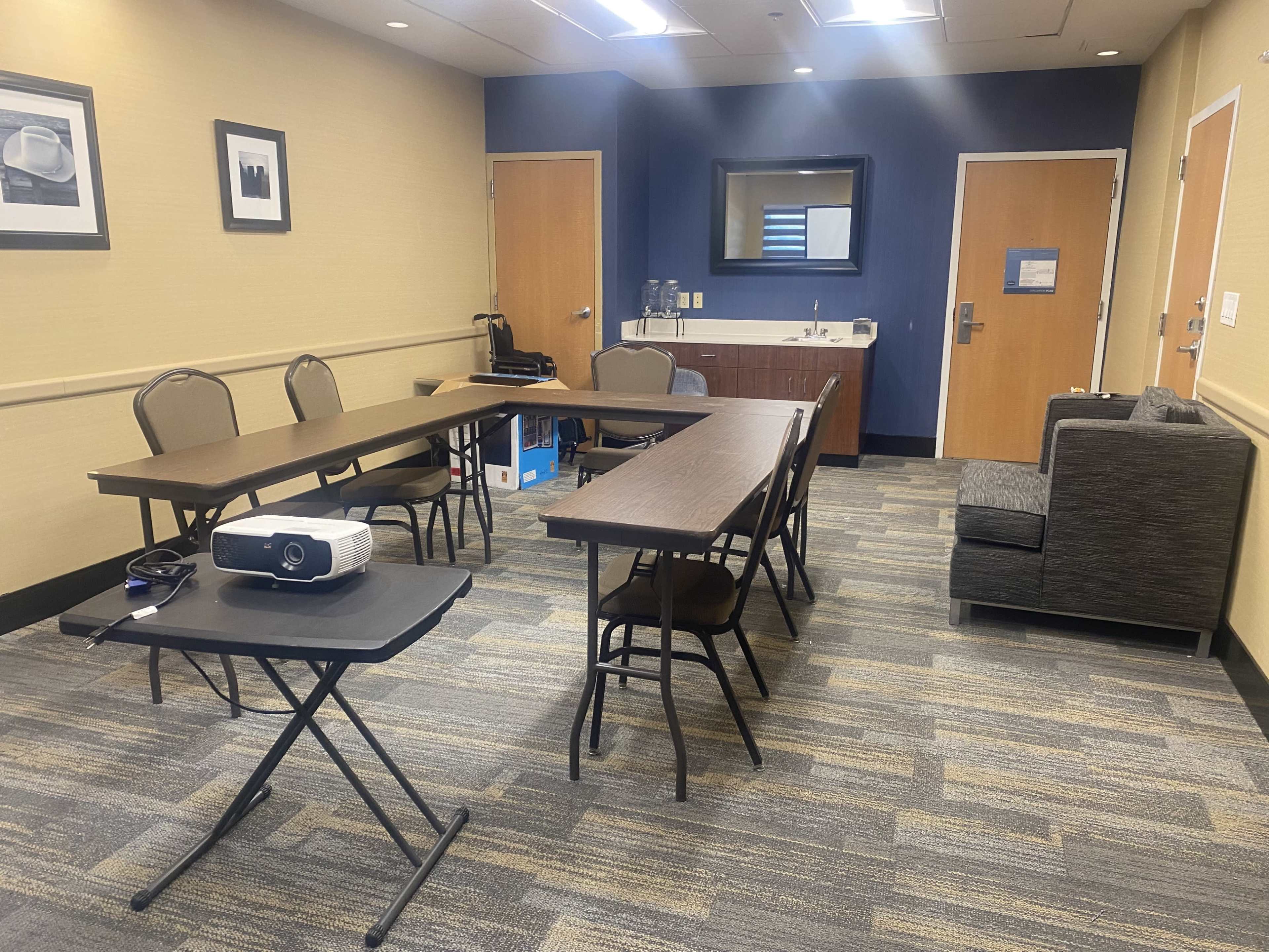 A small conference room with two long tables arranged in an L-shape, a projector on a cart, and a sofa in one corner.