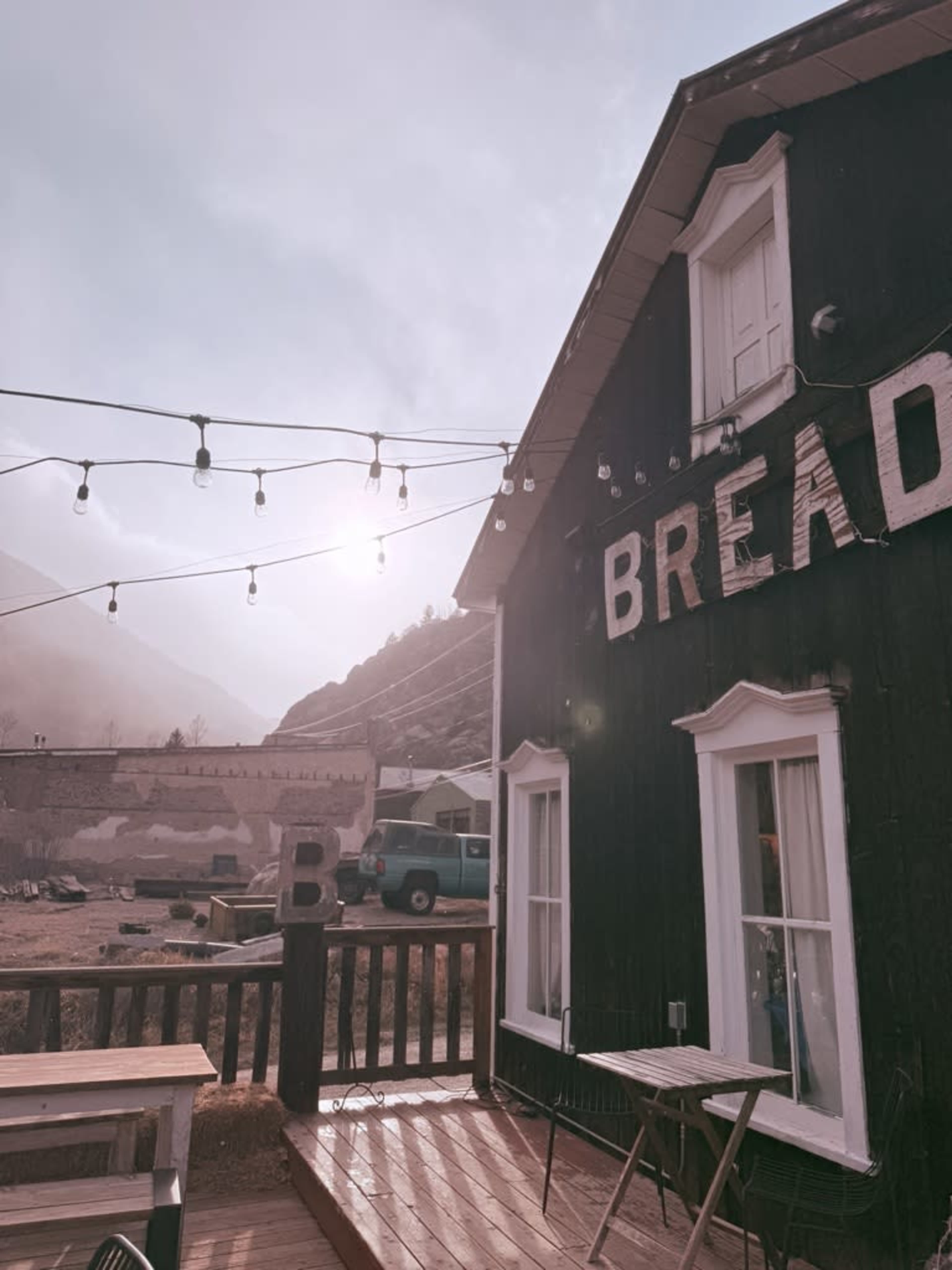 A wooden building with a sign that reads "BREAD" is partially obscured by fog, with string lights overhead and mountains in the background.