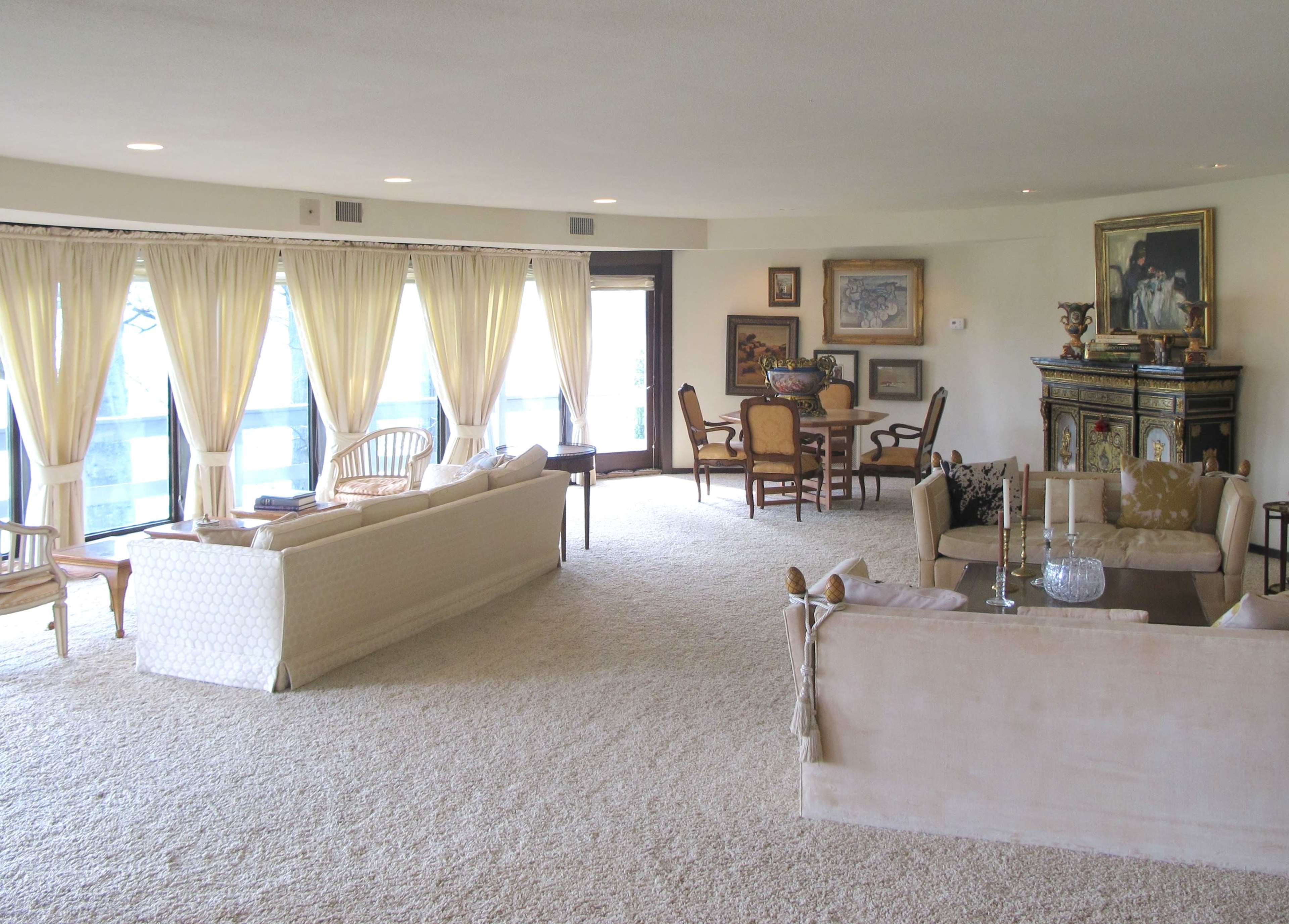 The image shows a spacious living room with large windows, white curtains, and a mix of seating arrangements including sofas and chairs, along with decorative art pieces on the walls.