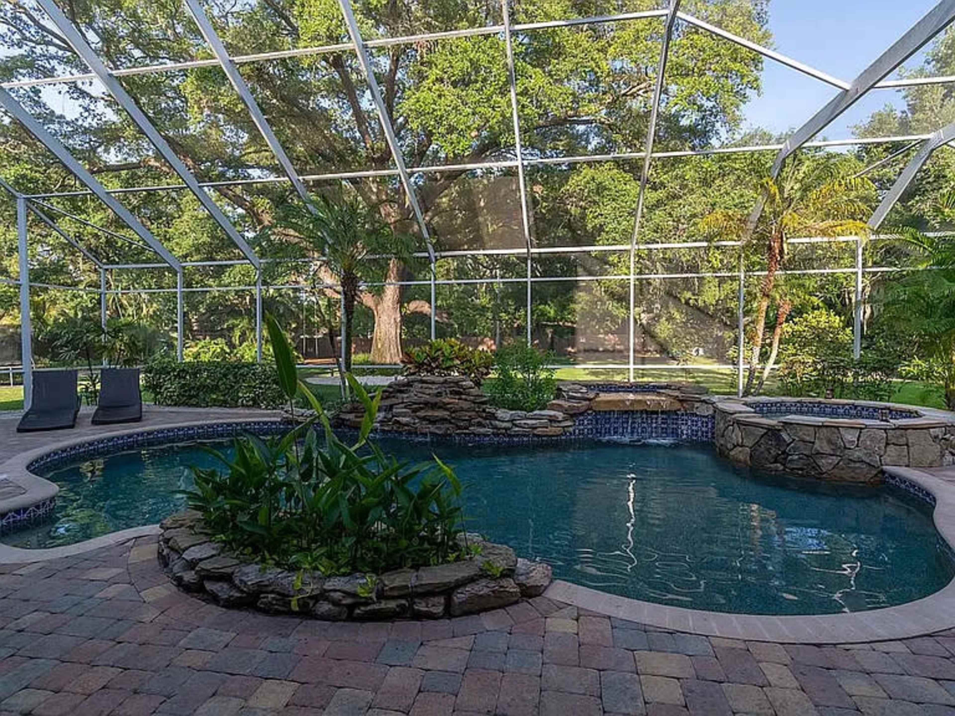 The image shows a backyard pool enclosed by a metal screen, featuring a rock formation and surrounding greenery.
