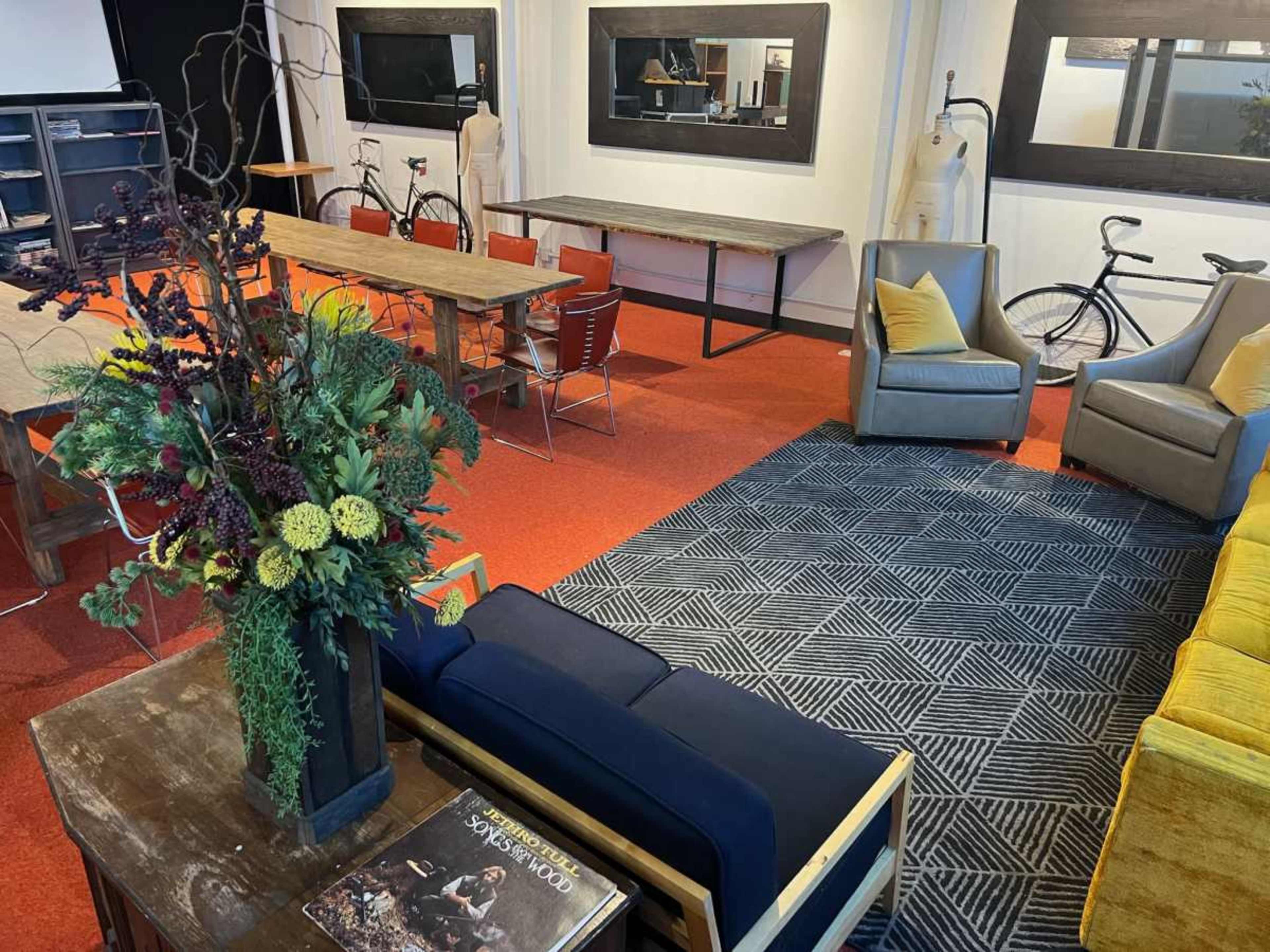 The image shows a cozy, modern office space featuring a mix of seating areas, tables, and decorative plants.