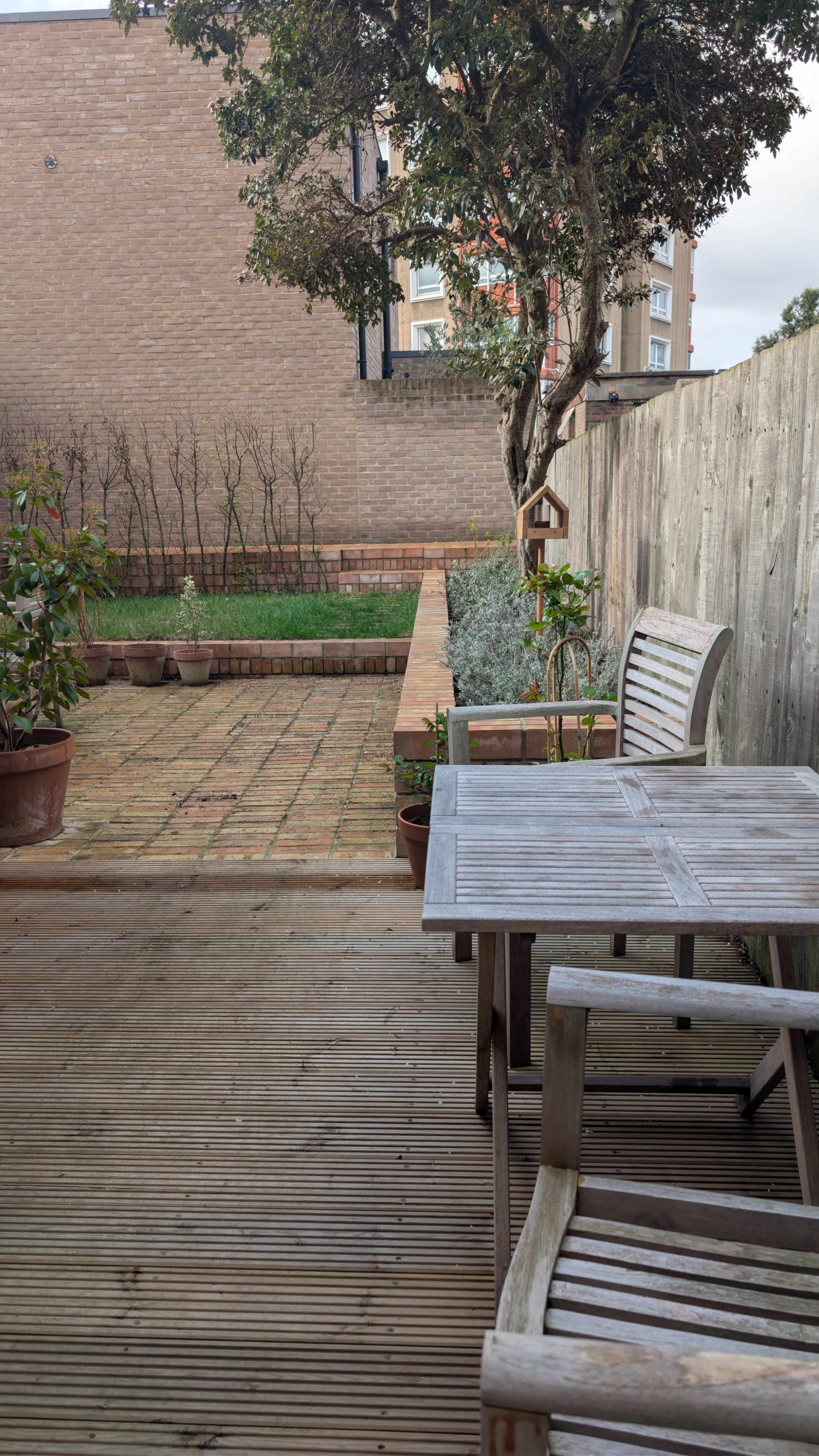 The image shows a wooden deck with a table and chairs overlooking a brick-paved garden area bordered by a wooden fence and containing potted plants.