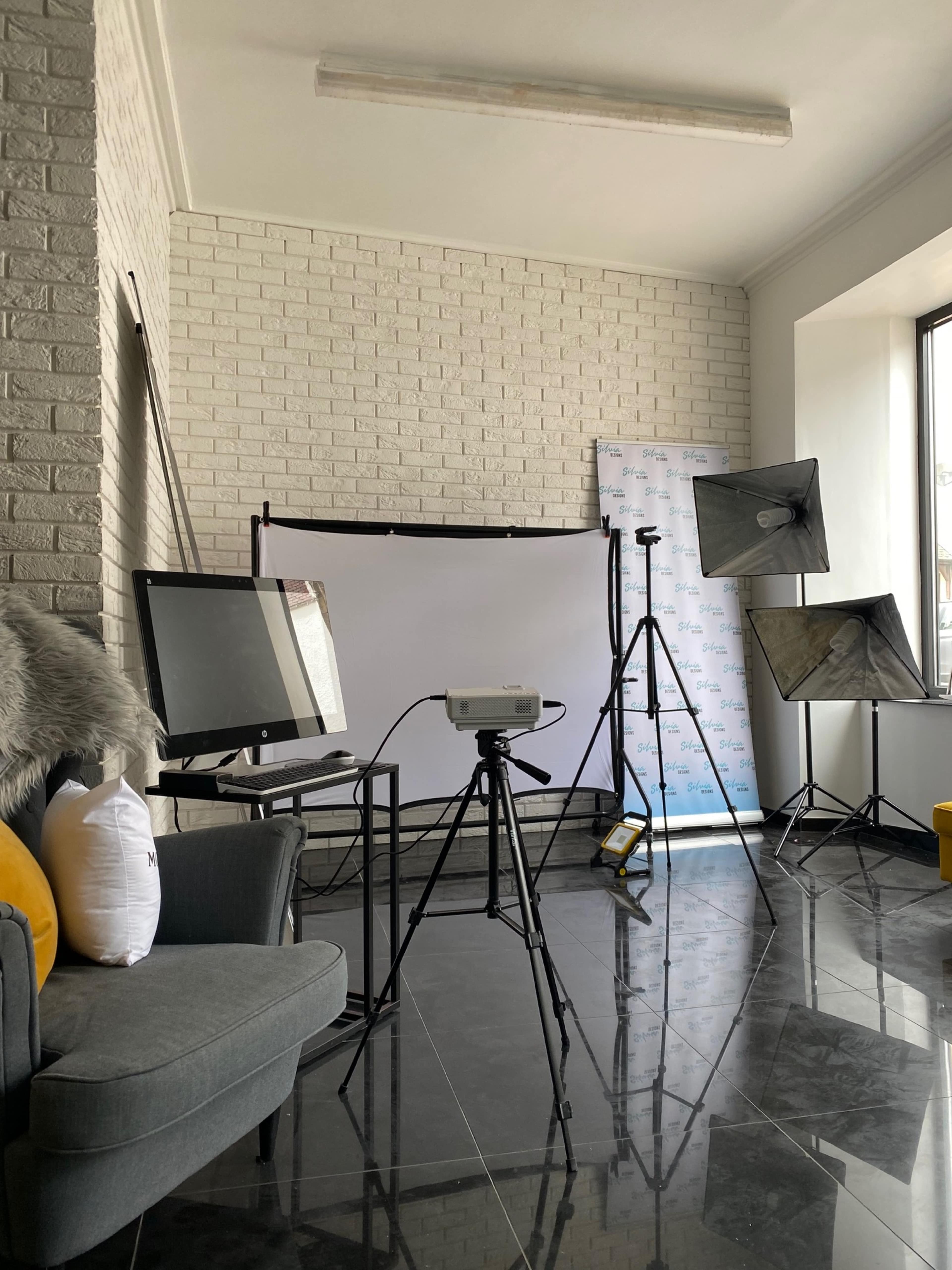 A room with a gray couch, a projector on a tripod, and two lighting setups in front of a white backdrop.