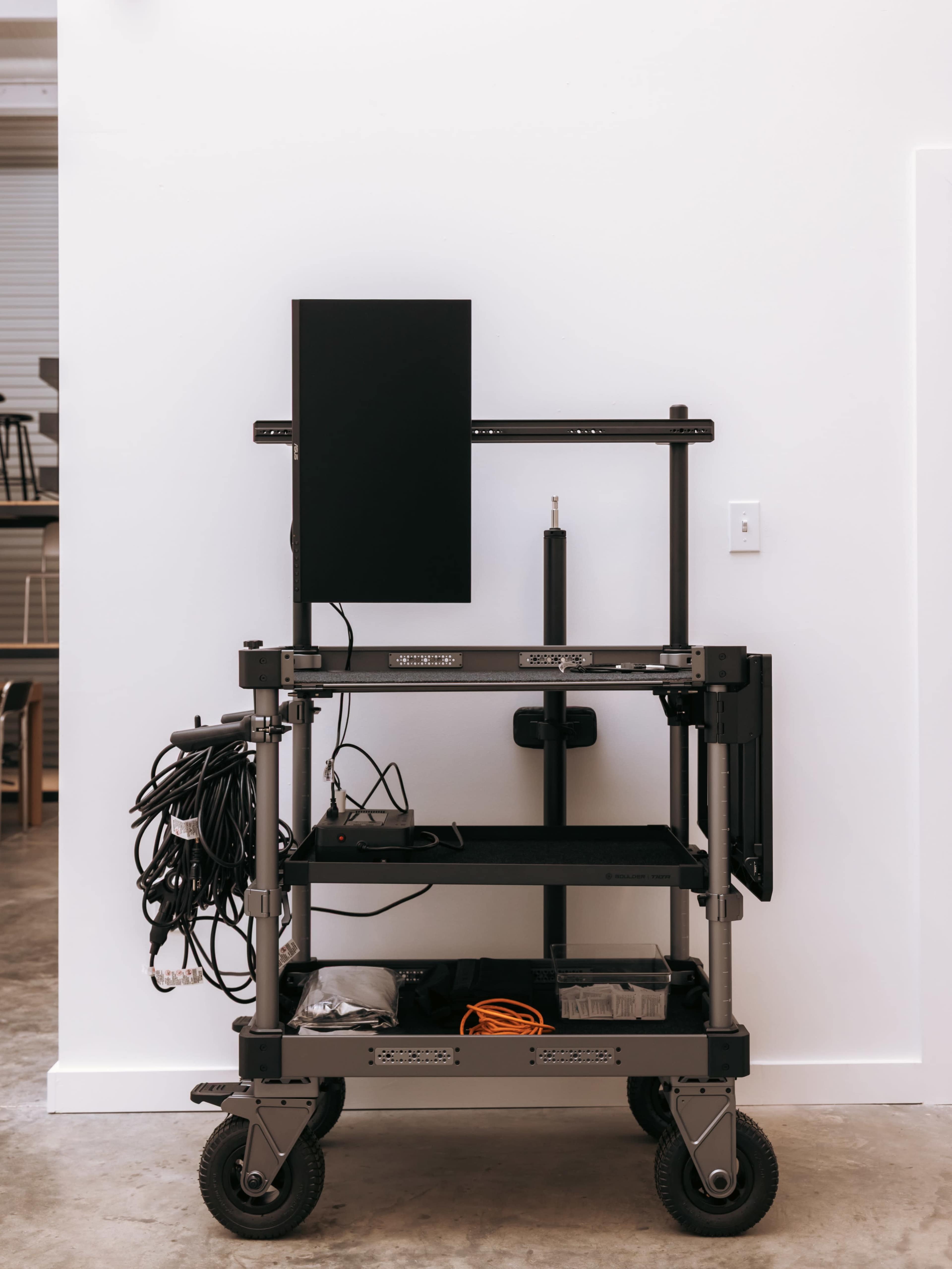 A mobile cart is positioned against a white wall, featuring a monitor on top and various wires organized on the shelves below.