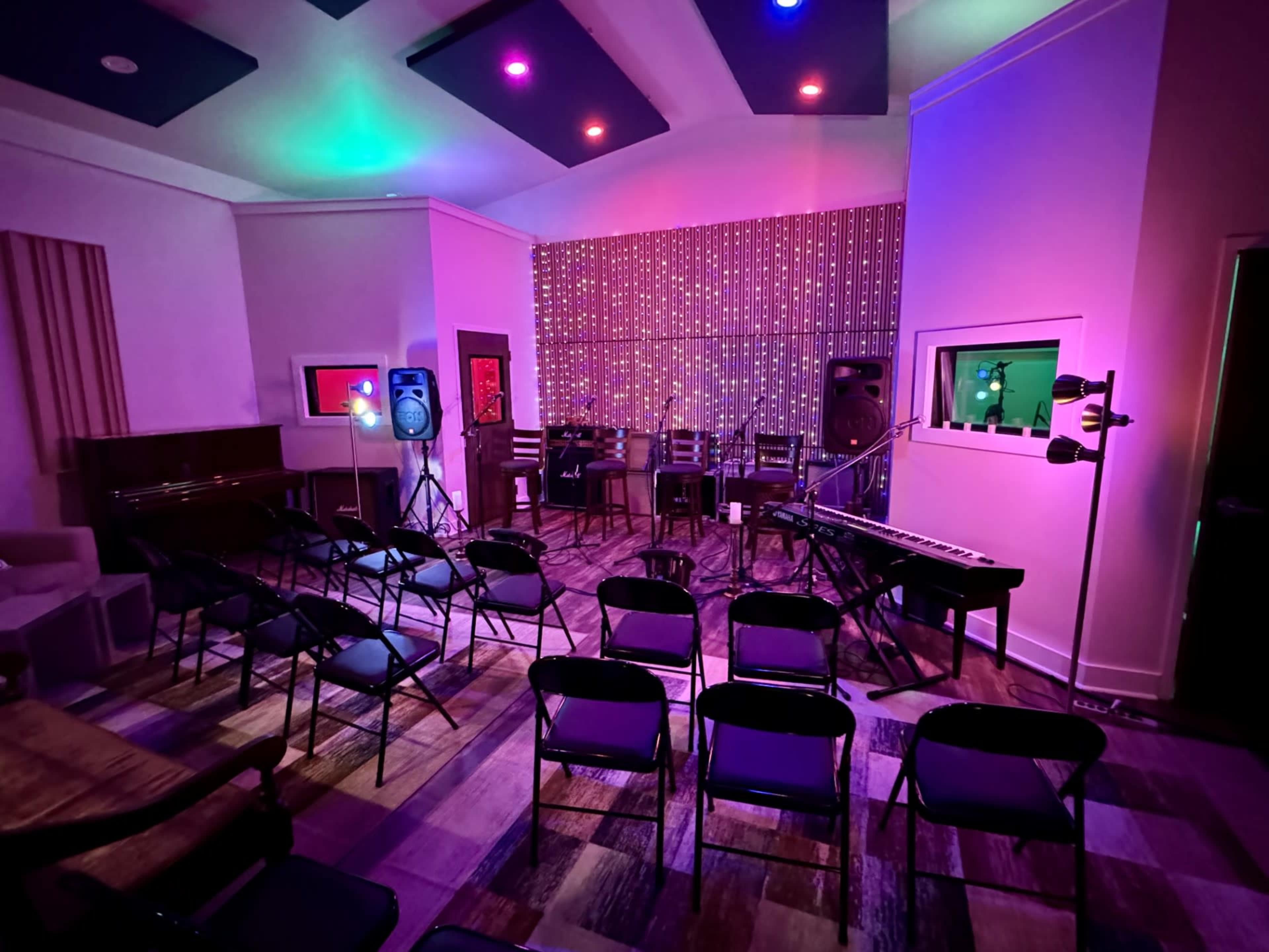 The image depicts a music studio set up for a performance, featuring chairs arranged in front of a stage with colorful lighting and sound equipment.