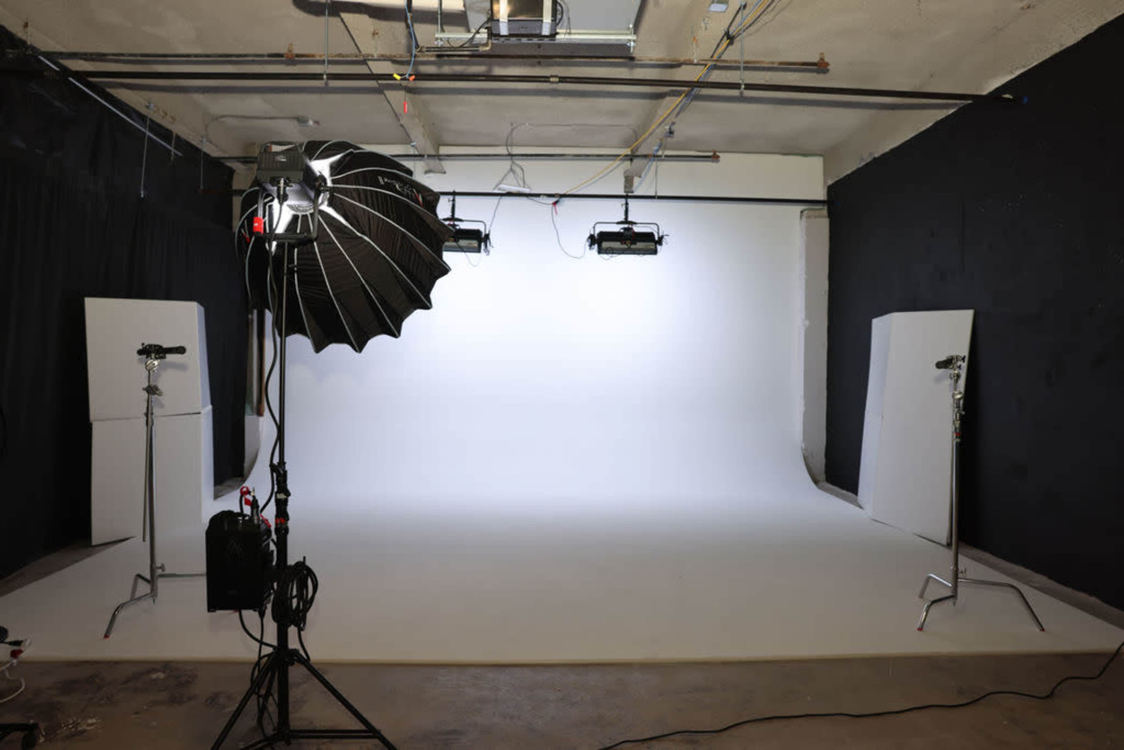 The image shows a photography studio with a plain white backdrop and several lighting setups.