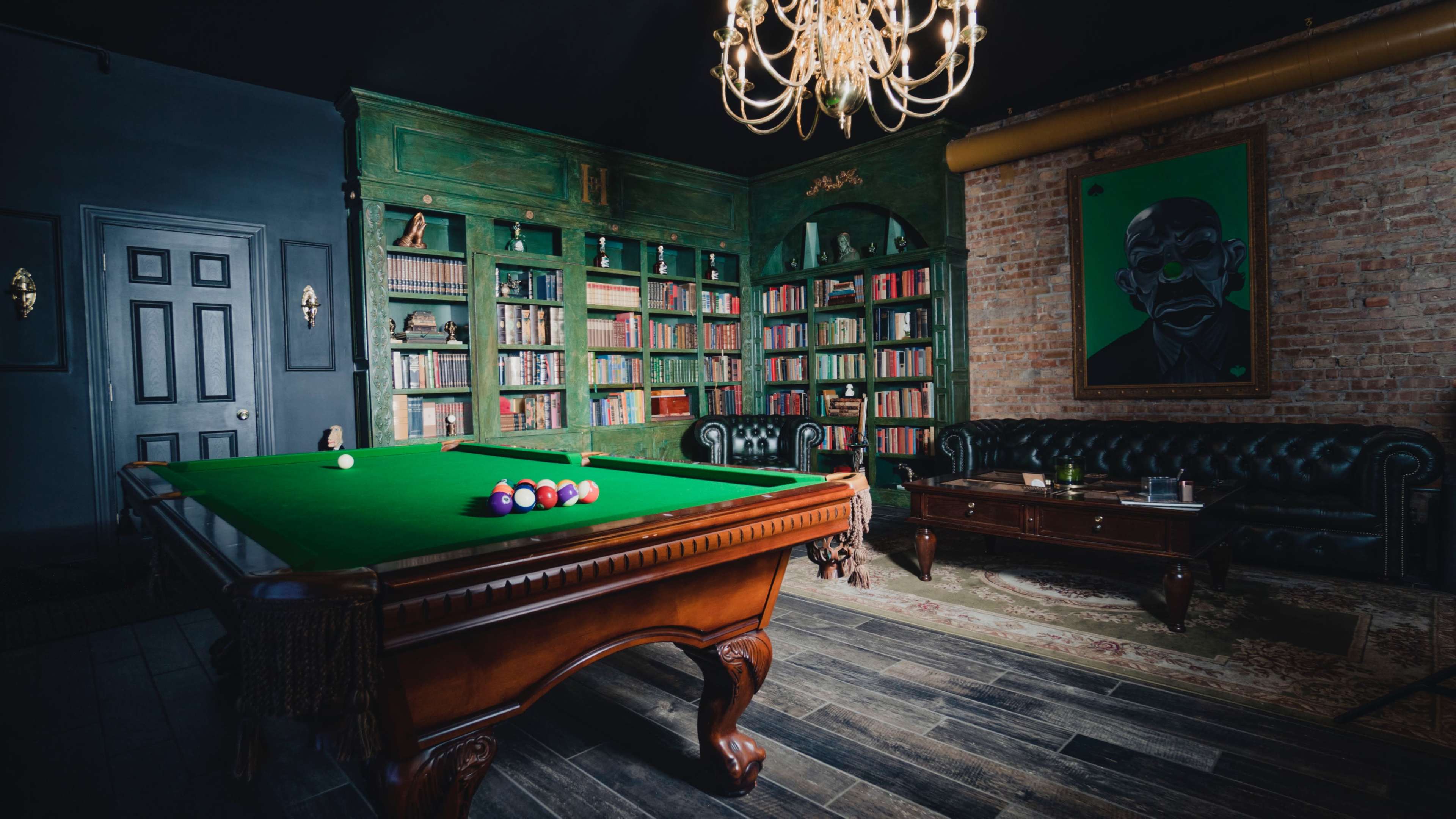 A room with a green pool table in the center, surrounded by bookshelves filled with books, a leather sofa, and a chandelier overhead.