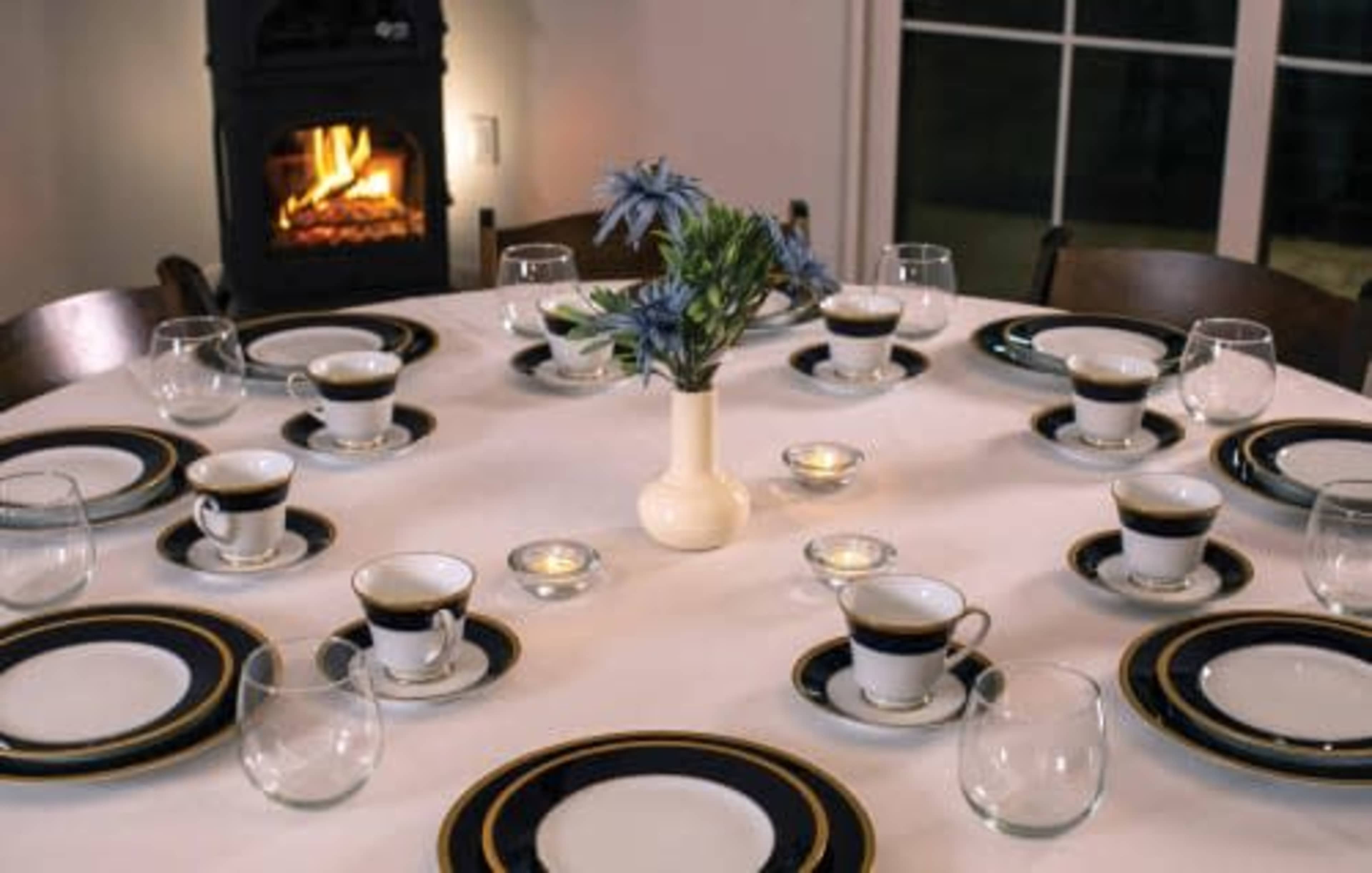 A round dining table is set with plates, cups, and glasses, centered by a vase of flowers and illuminated by candles, with a fireplace visible in the background.