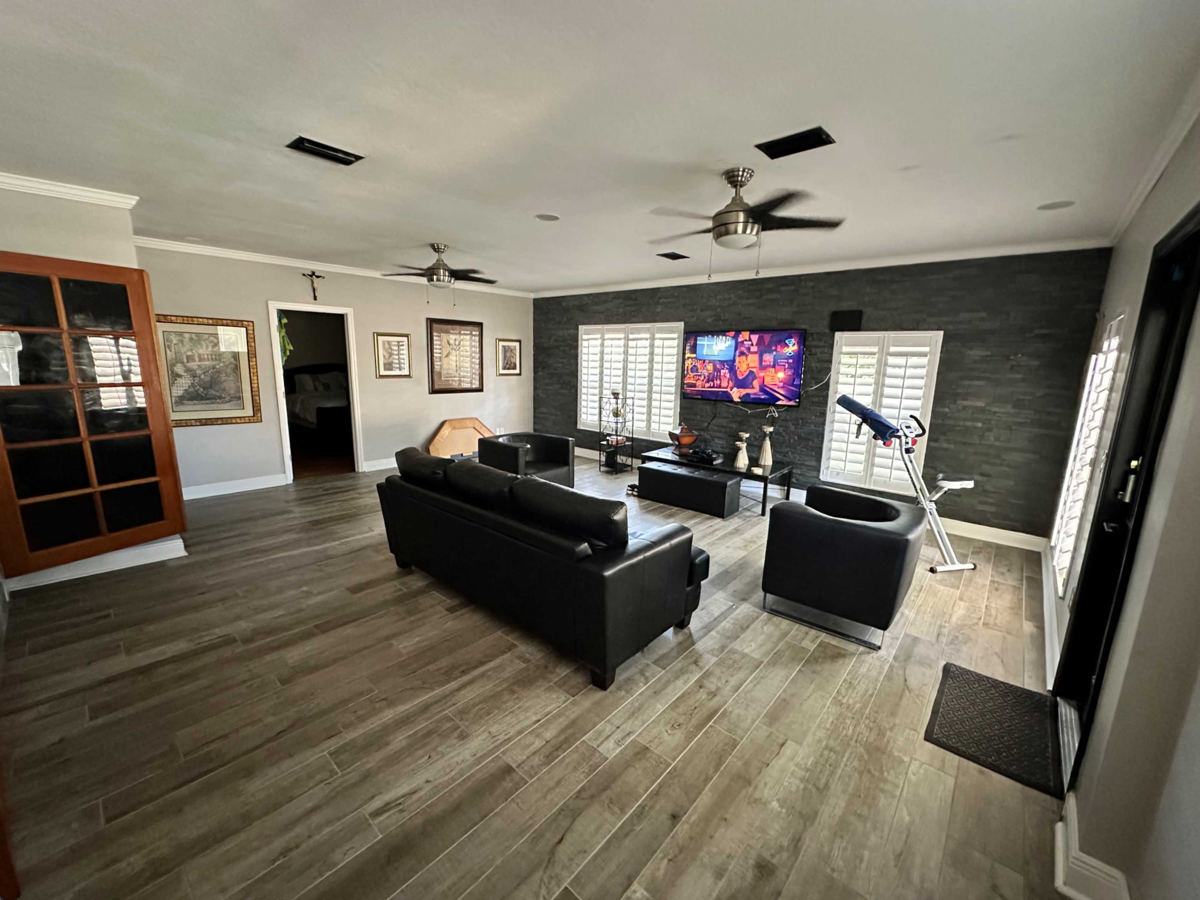 A living room with black leather furniture, a wall-mounted television, and light-colored wooden flooring.