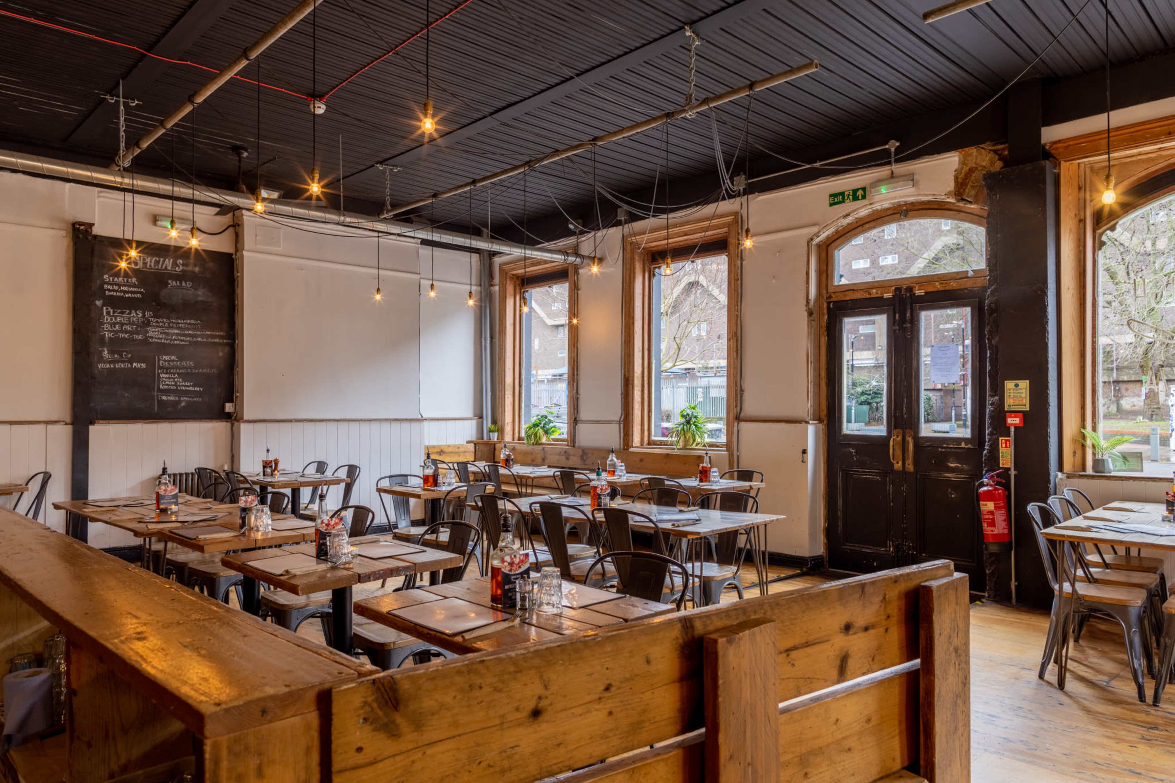The interior of a restaurant features wooden tables and chairs arranged in a spacious layout, with large windows allowing natural light to enter.