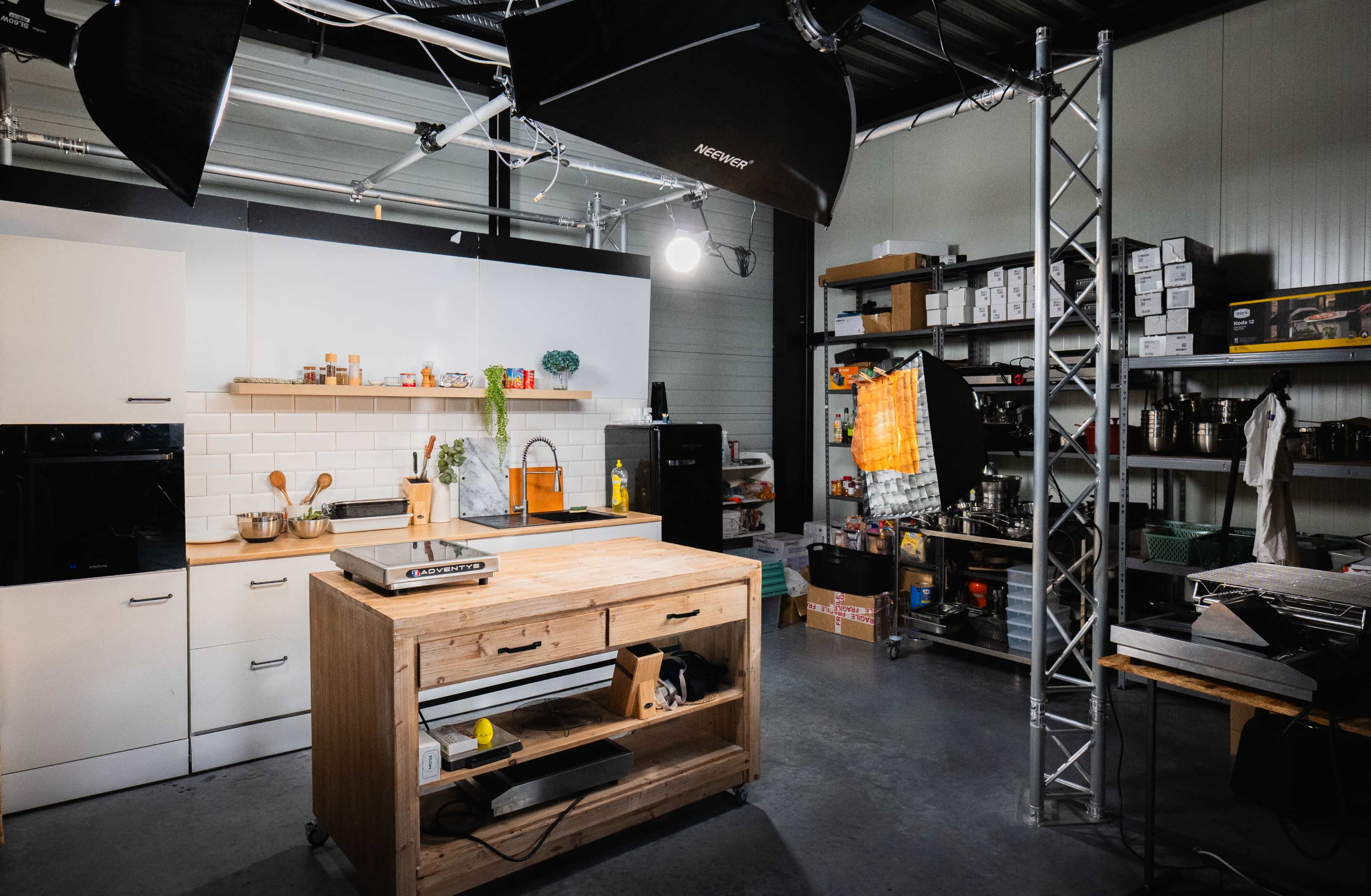 The image shows a modern kitchen studio with white cabinets, an island, and various kitchen tools and props arranged neatly on shelves and counters.