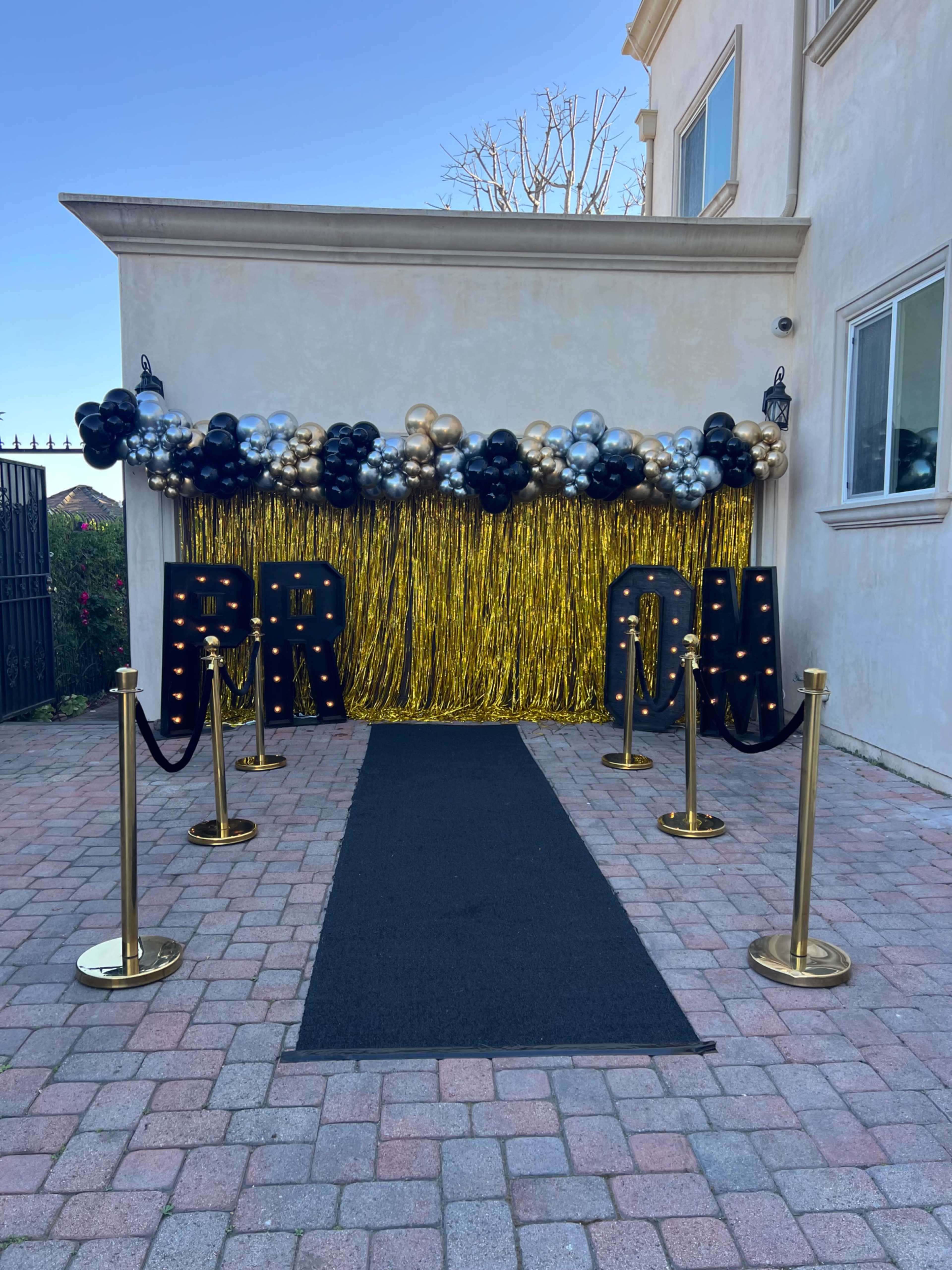 The image shows a decorated entrance for a prom event, featuring a black carpet, gold stanchions, large letters spelling "PROM," and a backdrop of balloons and shiny gold streamers.