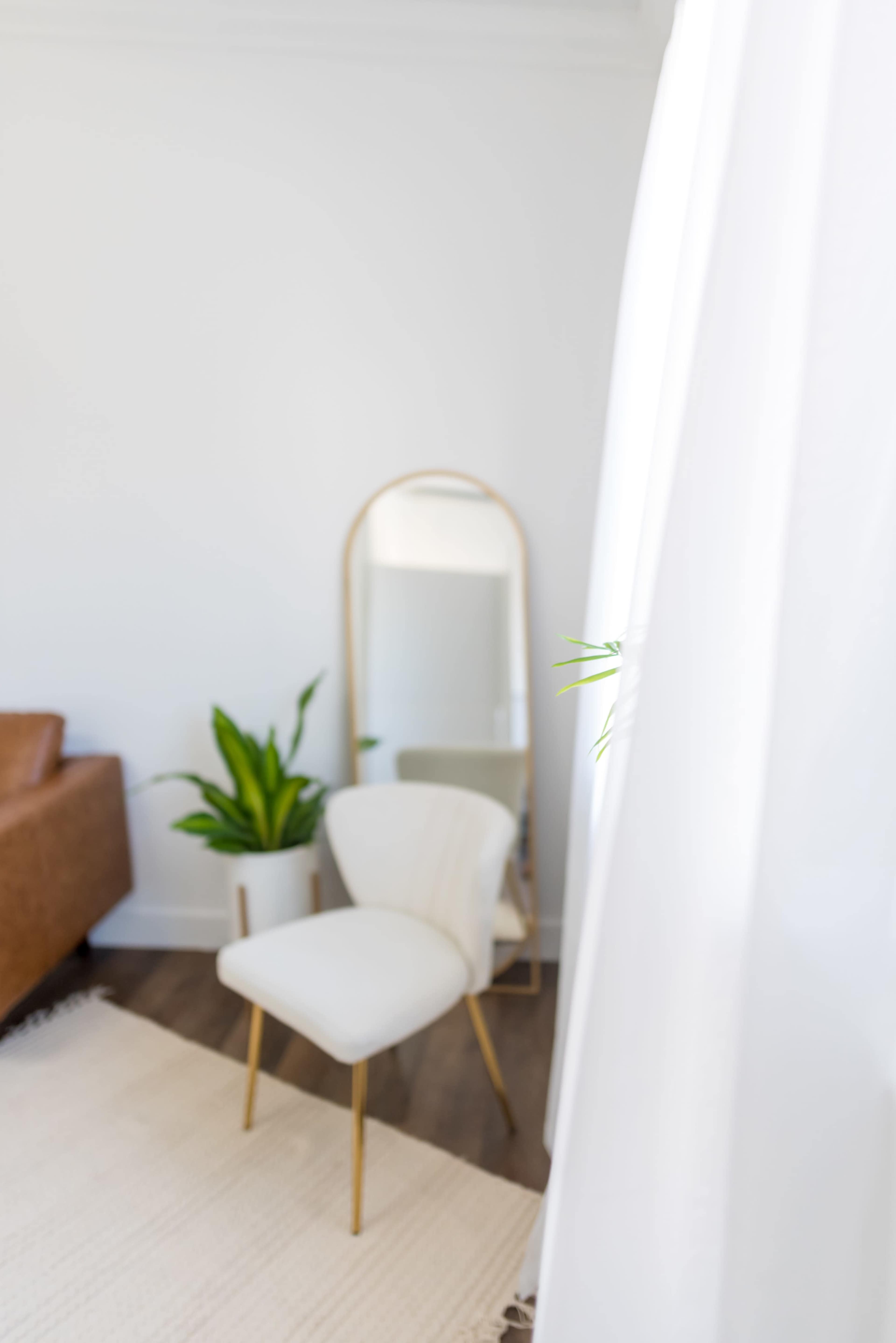 A white chair with gold legs is positioned next to a mirror and a potted plant in a bright room.