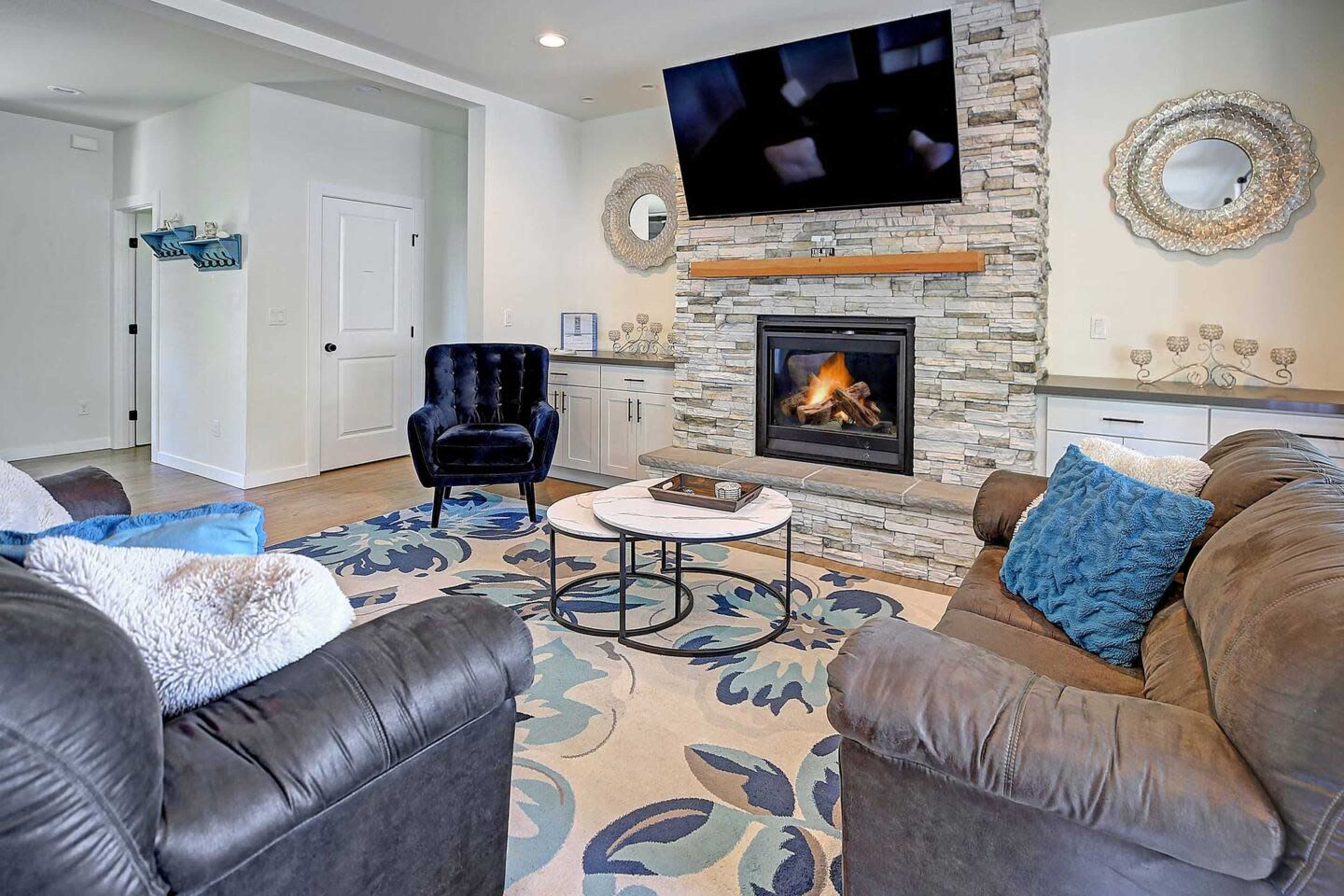The image shows a cozy living room featuring a stone fireplace, a mounted television, and two sofas arranged around a circular coffee table on a floral-patterned rug.