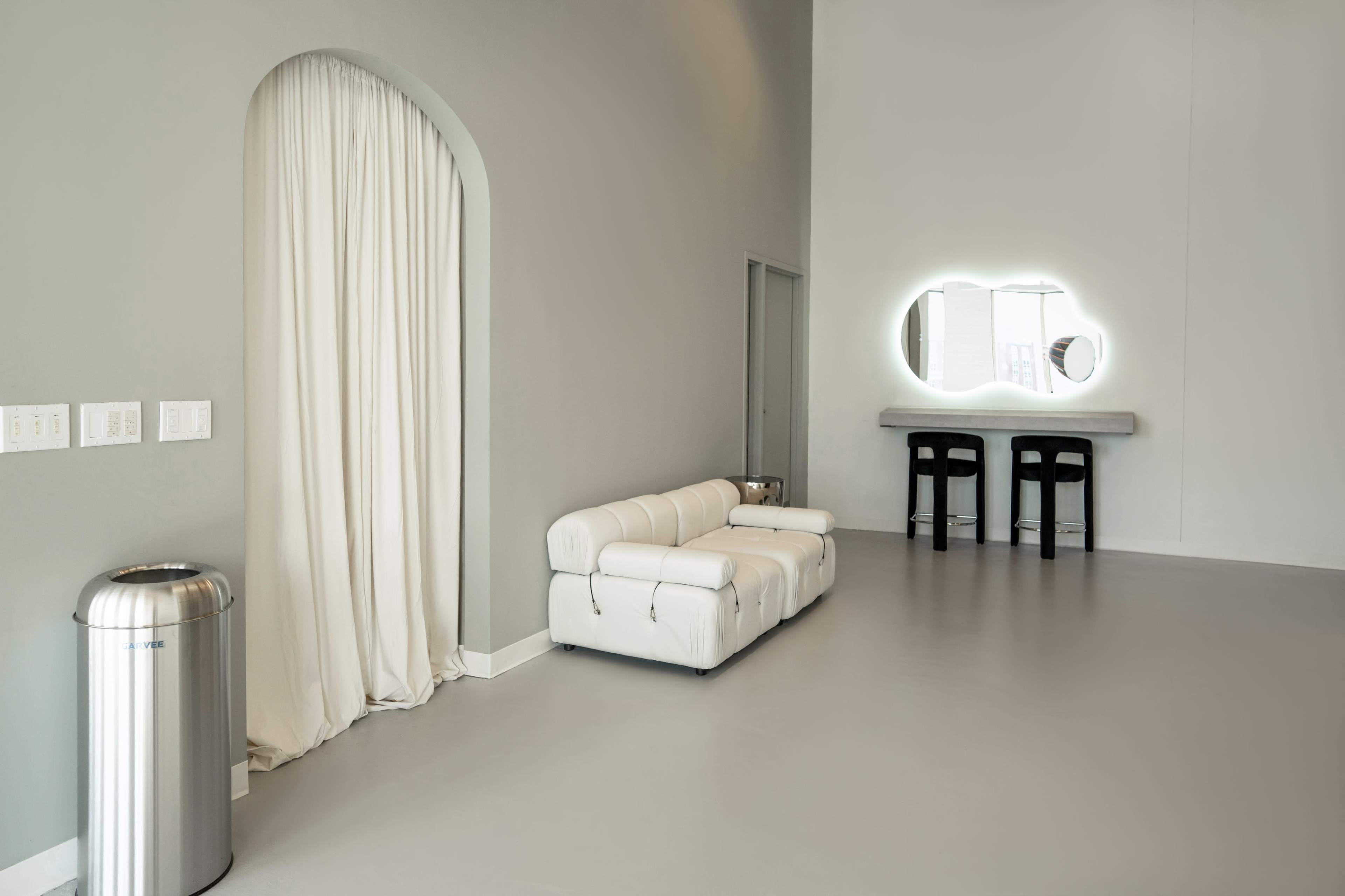 The image shows a spacious, minimalist interior featuring a white sofa, a mirror with ambient lighting, and a small table with two stools.