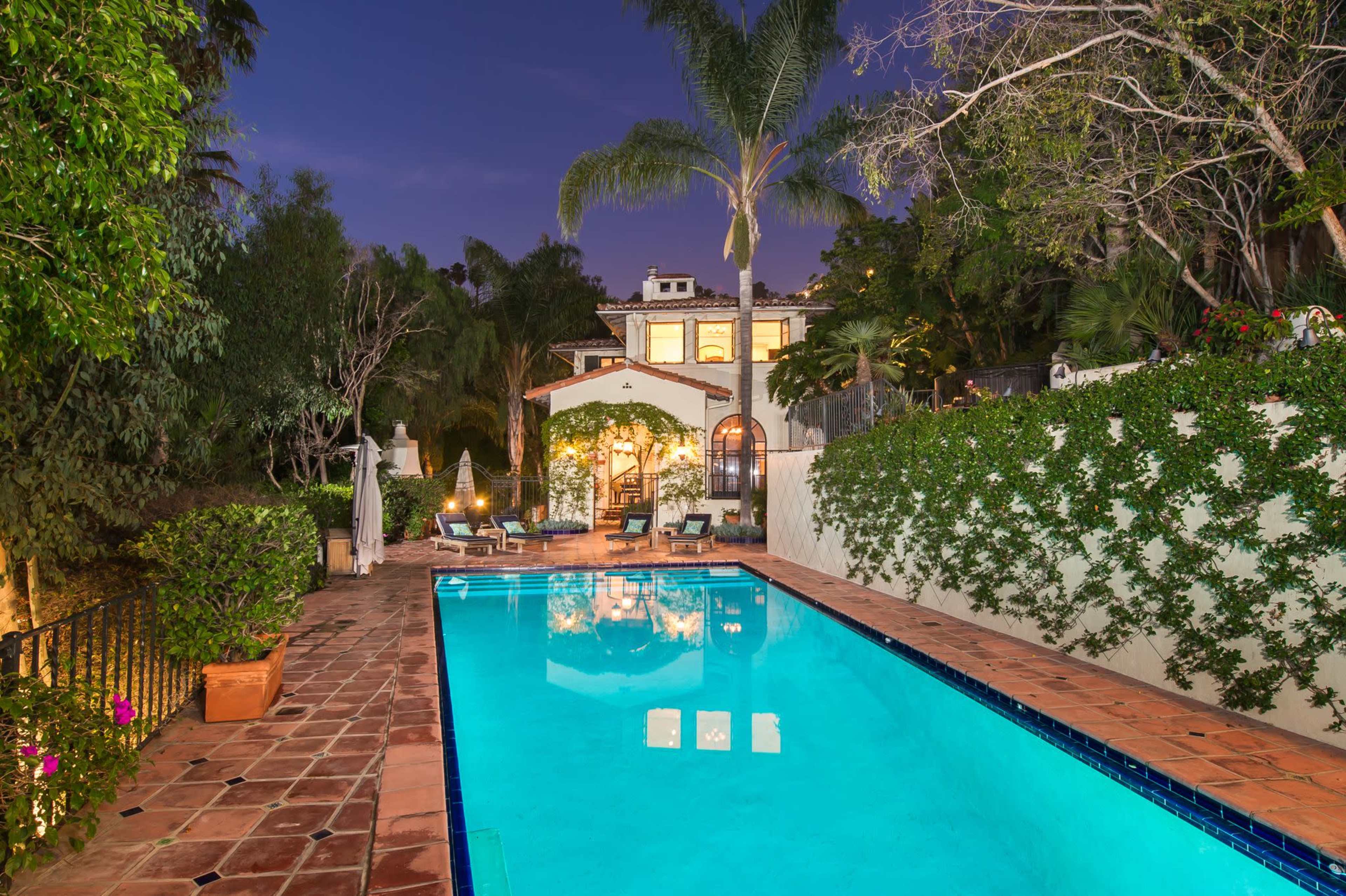 A well-lit pool reflects the surrounding greenery and a two-story house at dusk.