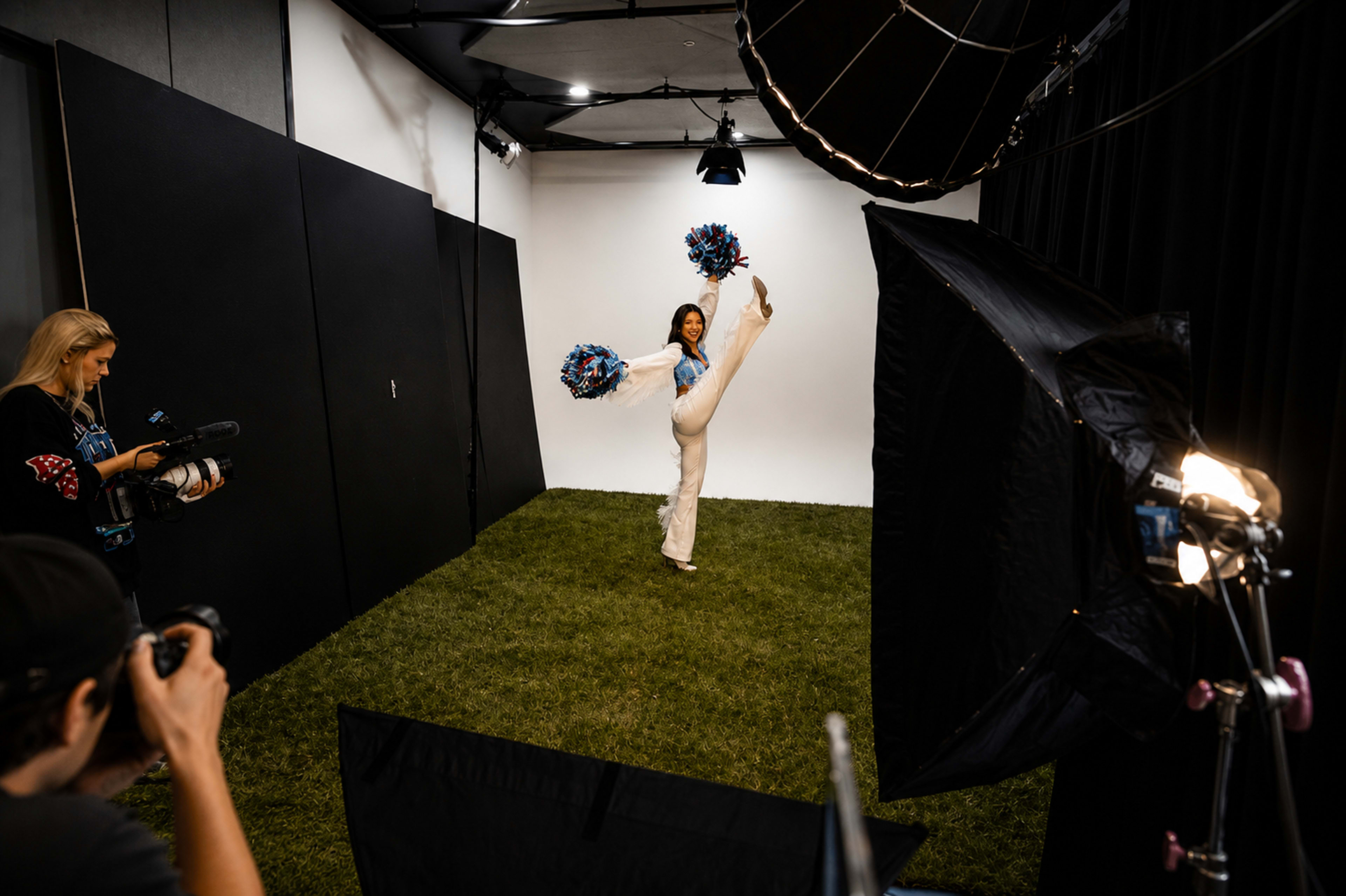A person in a cheerleading outfit performs a high kick while holding pom-poms in a photography studio with artificial turf flooring.