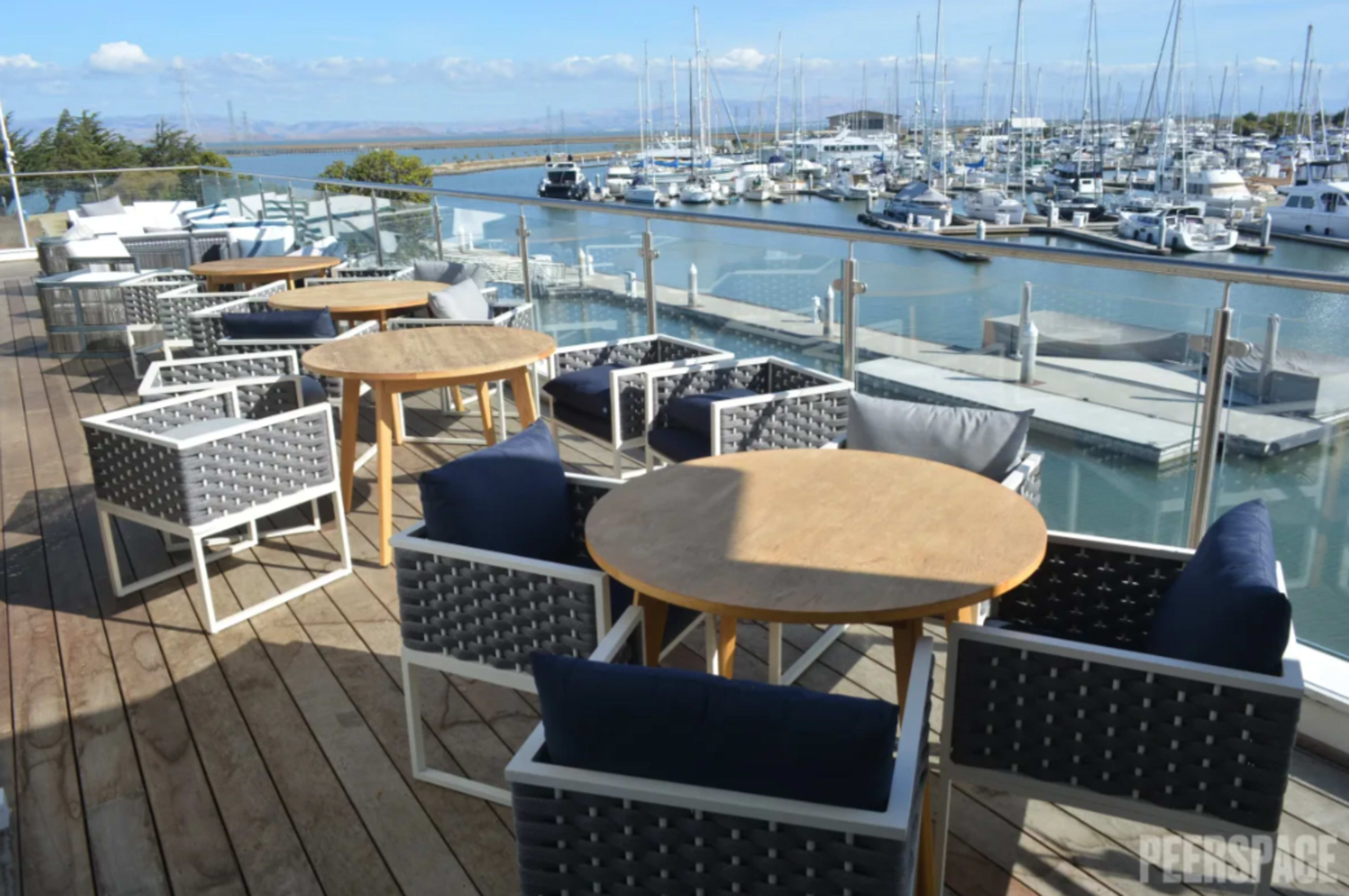 The image shows an outdoor terrace with wooden tables and modern chairs overlooking a marina filled with boats.