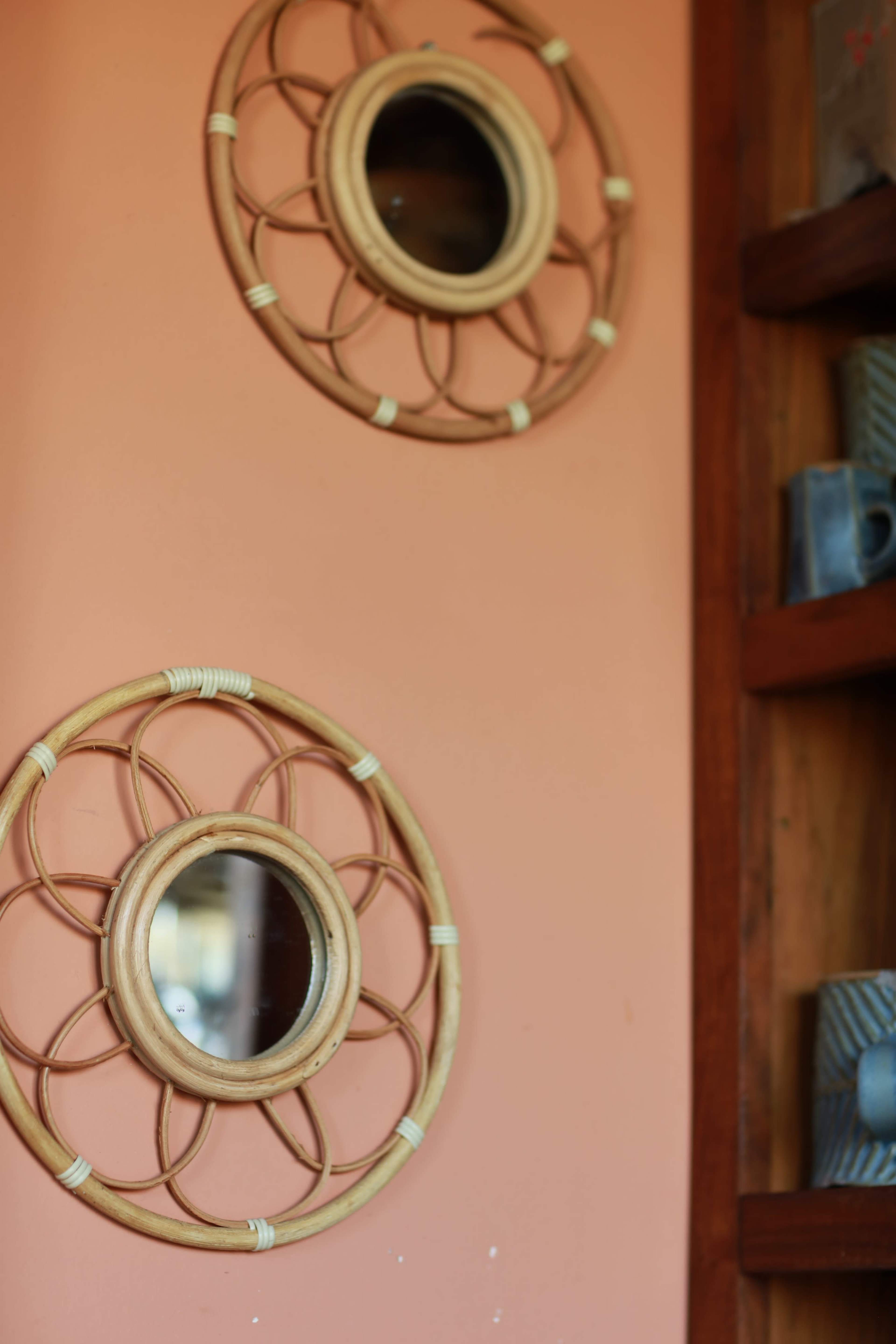Two round, woven mirrors are mounted against a peach-colored wall, with a wooden shelf displaying ceramic mugs nearby.