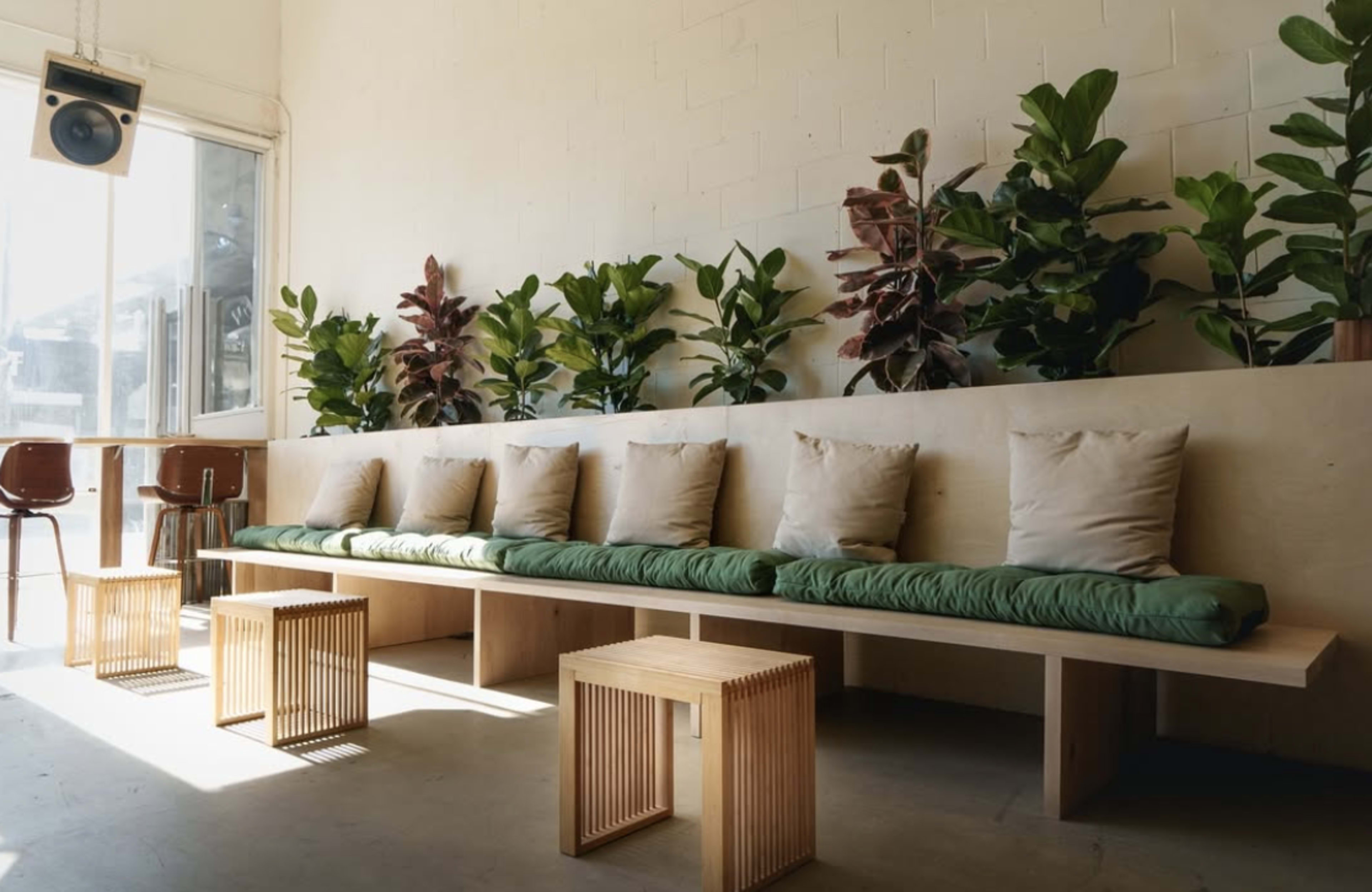 The image features a minimalist interior with a long bench, several cushions, and potted plants, alongside a small table and chairs.