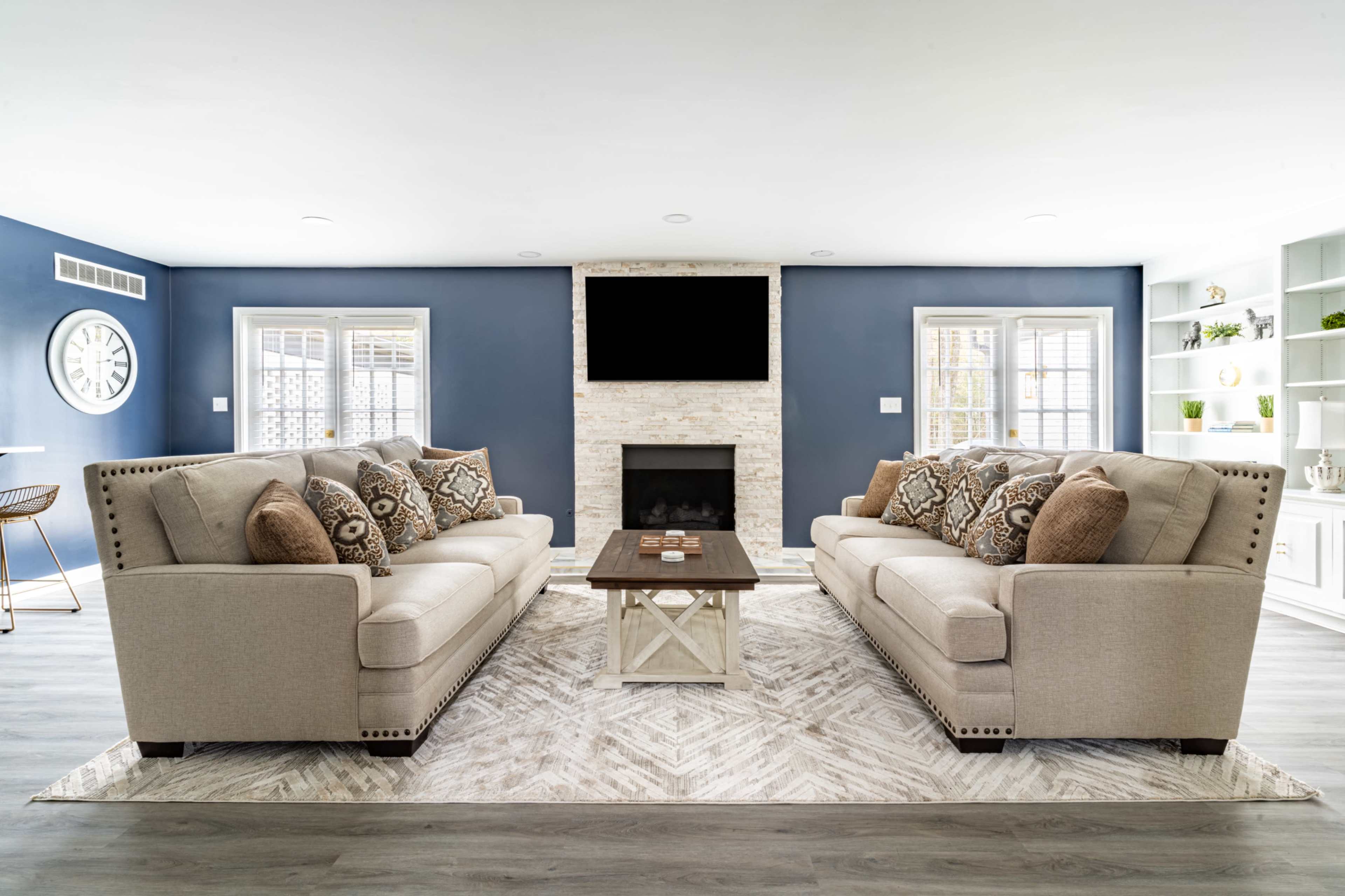 A modern living room features two beige sofas with decorative cushions facing a central coffee table on a patterned rug, framed by light-colored walls and a fireplace with a mounted television.
