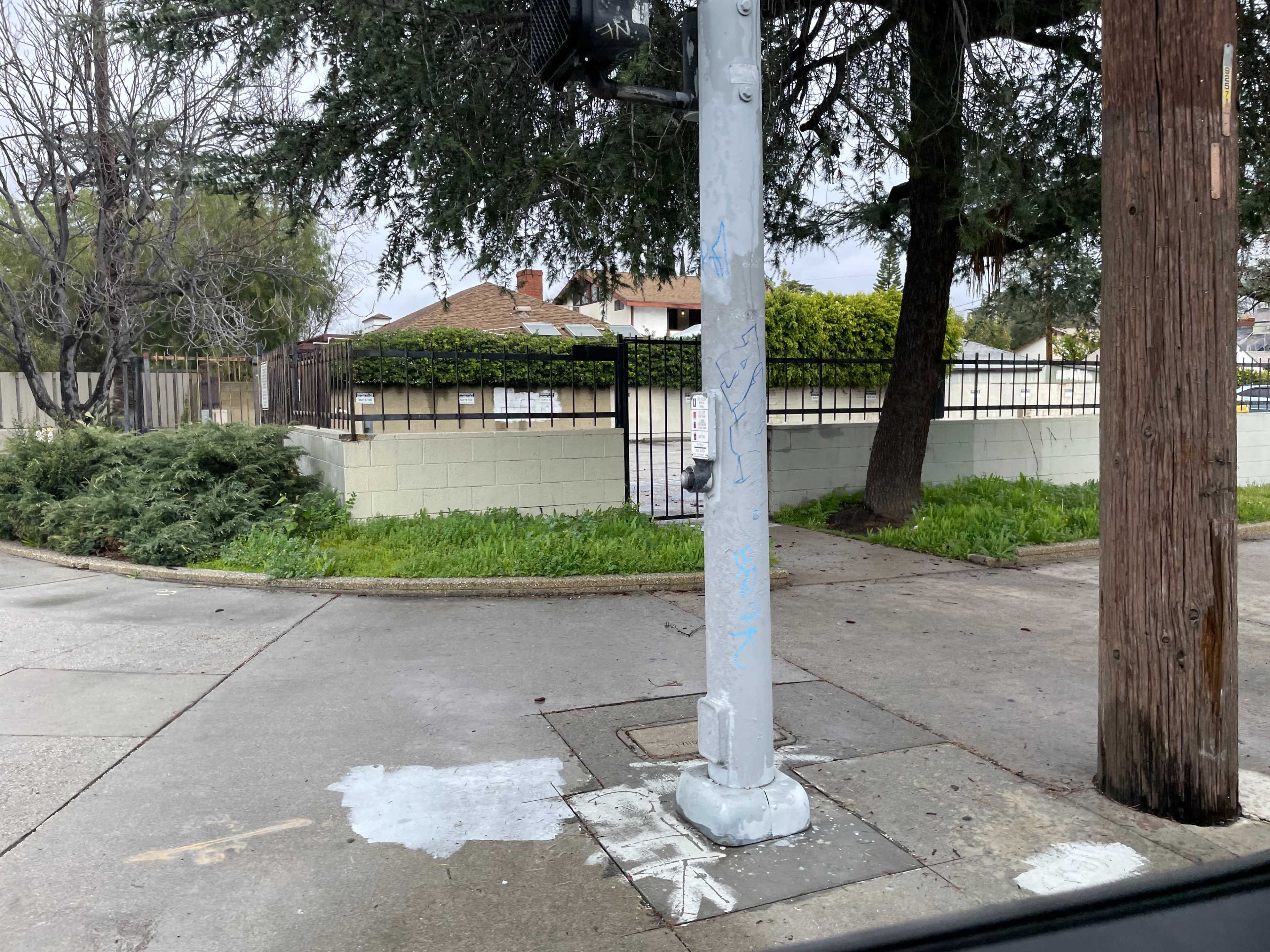 The image shows a street corner with a traffic signal pole, grassy areas, and a fenced residential property in the background.