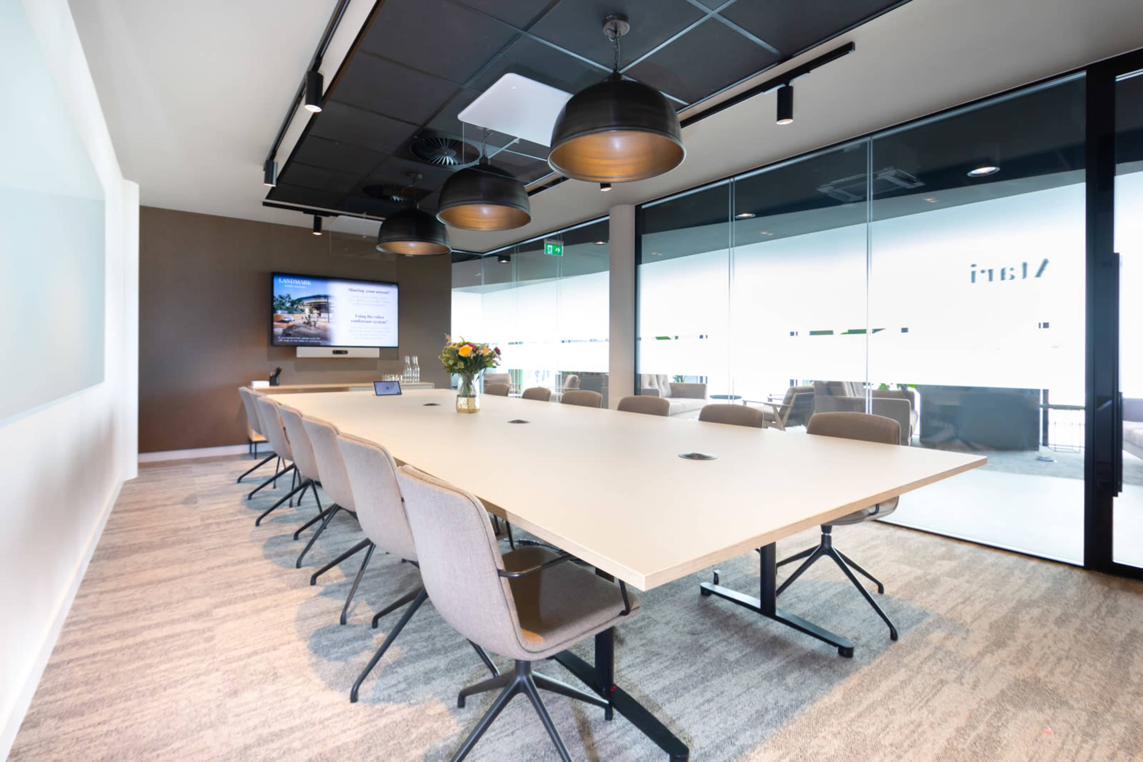 A modern conference room features a long table surrounded by gray chairs, with a large screen on the wall and windows displaying an outdoor view.