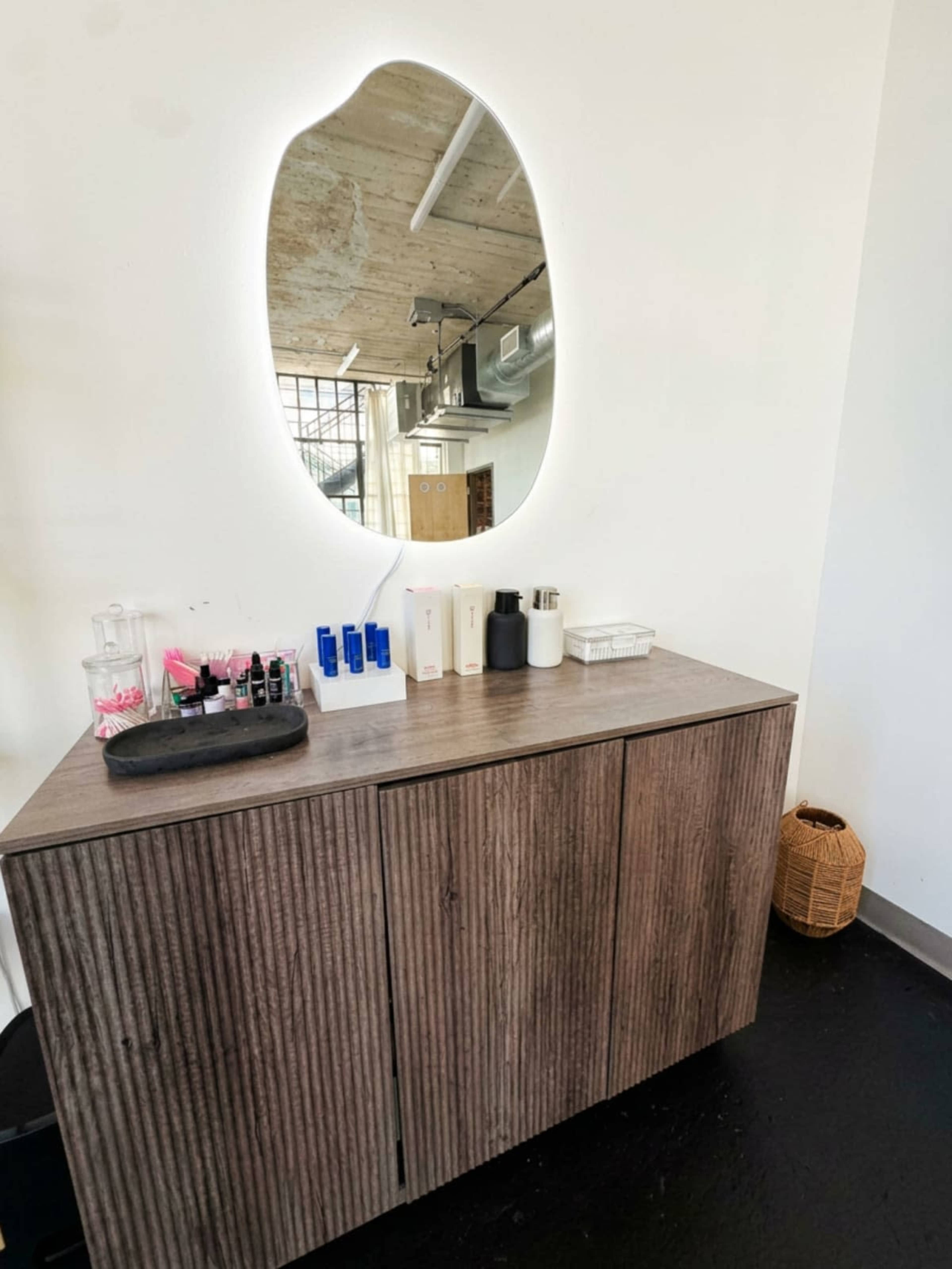 The image shows a wooden cabinet beneath an oval mirror, with various skincare products arranged on top, and a wicker basket nearby.