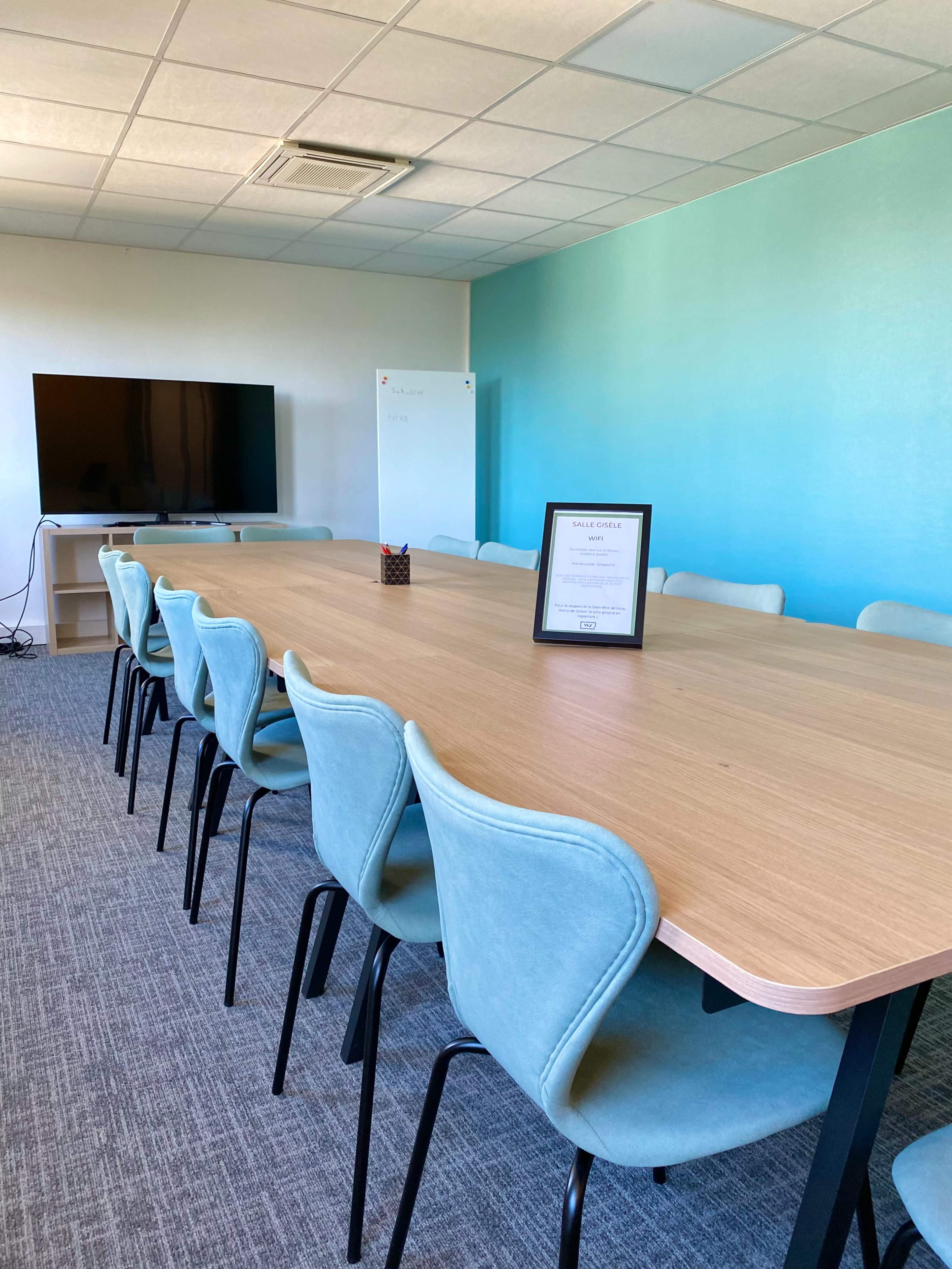 A long wooden table surrounded by light blue chairs is positioned in a bright meeting room with a television and a whiteboard on the walls.