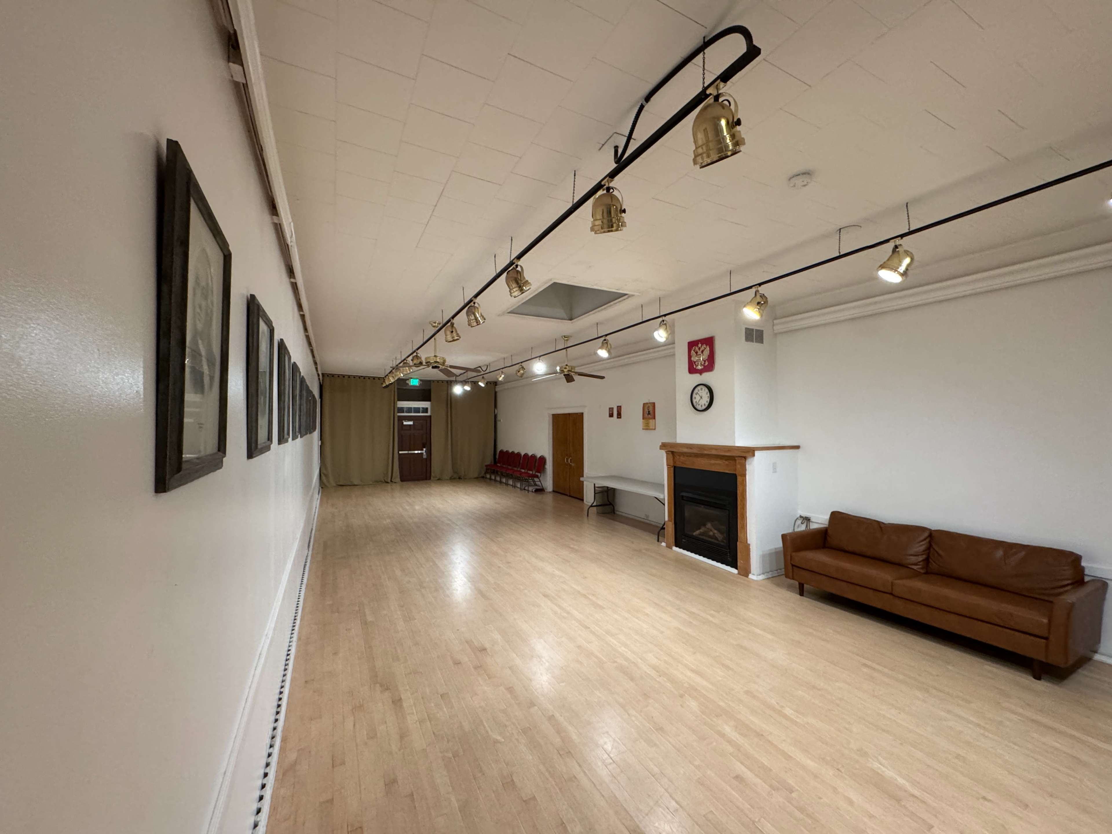 The image shows a spacious, well-lit room with a wooden floor, a long wall displaying framed pictures, a row of red chairs, and a brown sofa near a fireplace.