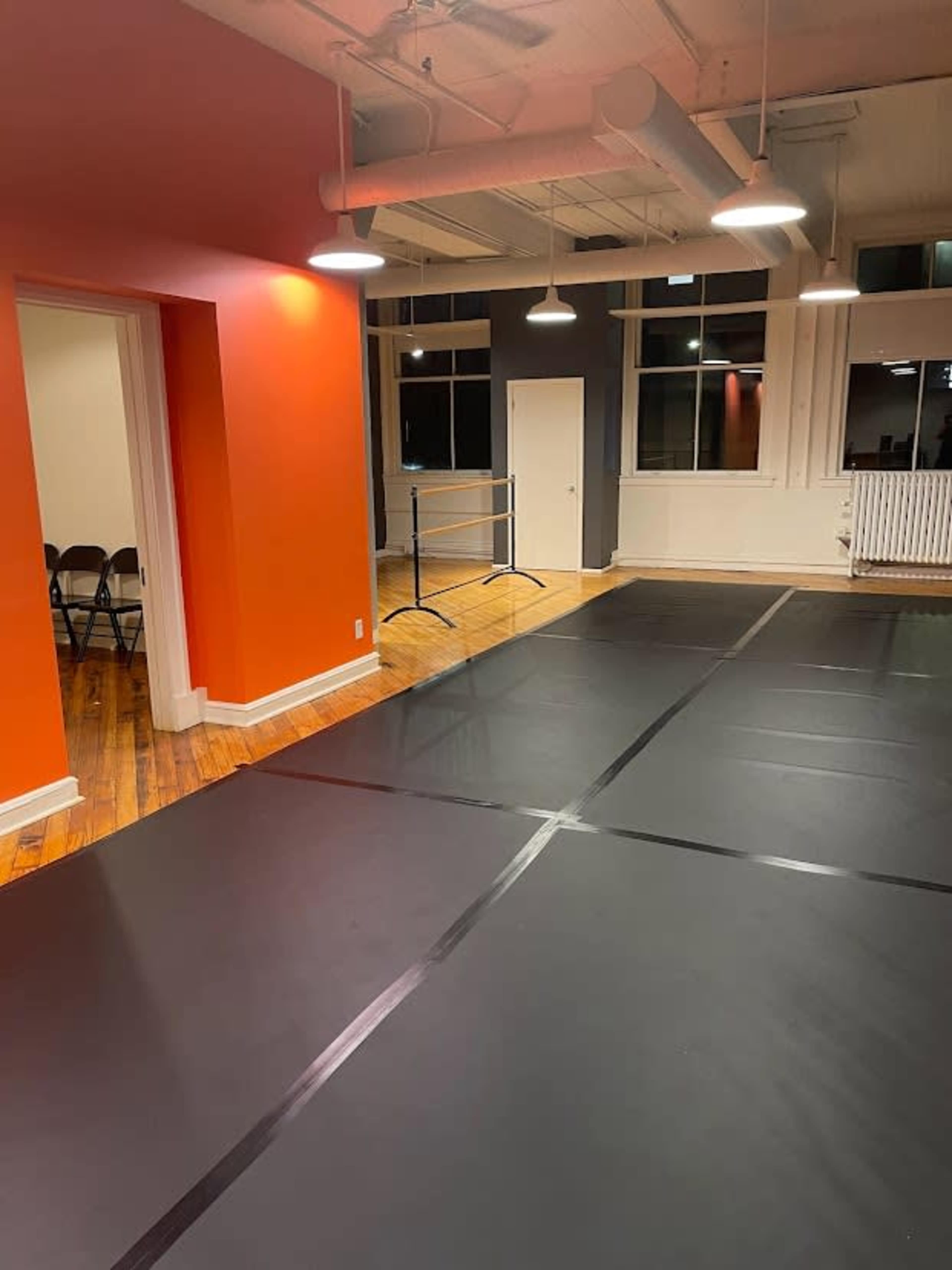 A spacious room with black mats on the floor, orange walls, and large windows illuminated by pendant lights.