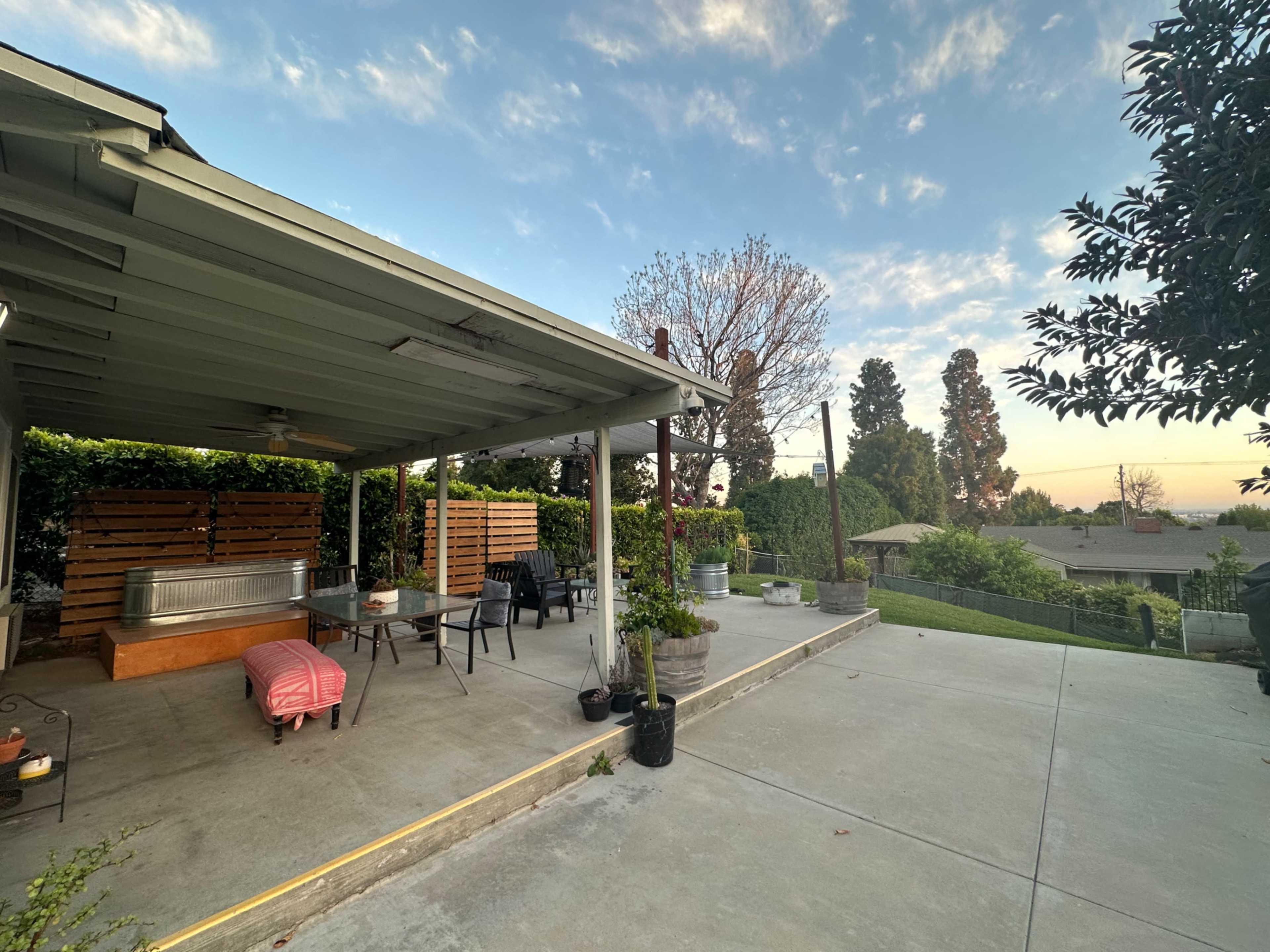 A patio area features a table and chairs under a roof, with a view of landscaped yards and trees in the background.