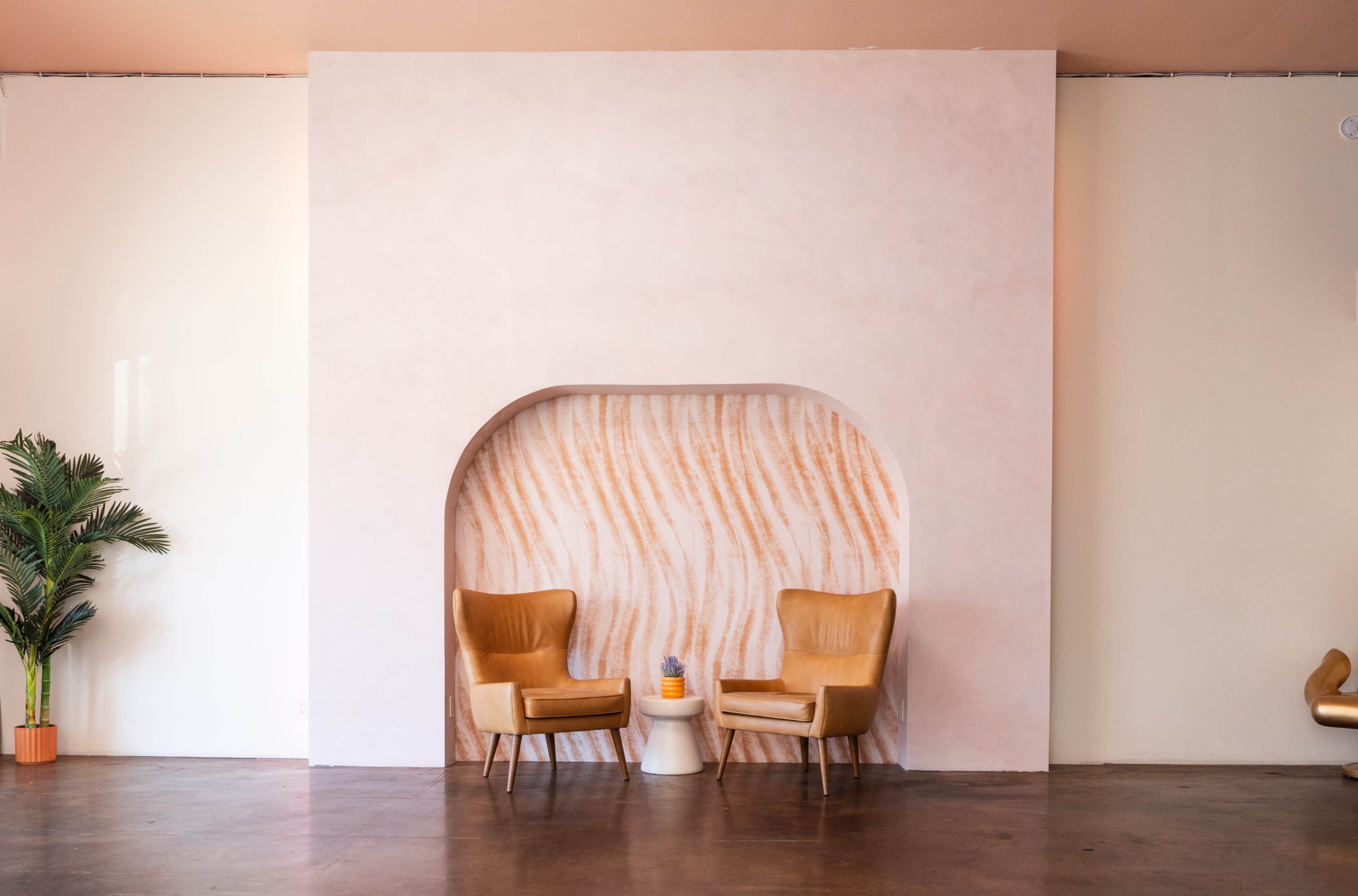 The image shows a minimalistic interior setting with two tan chairs and a small table in front of a curved wall featuring a subtle textured design, alongside a potted plant.
