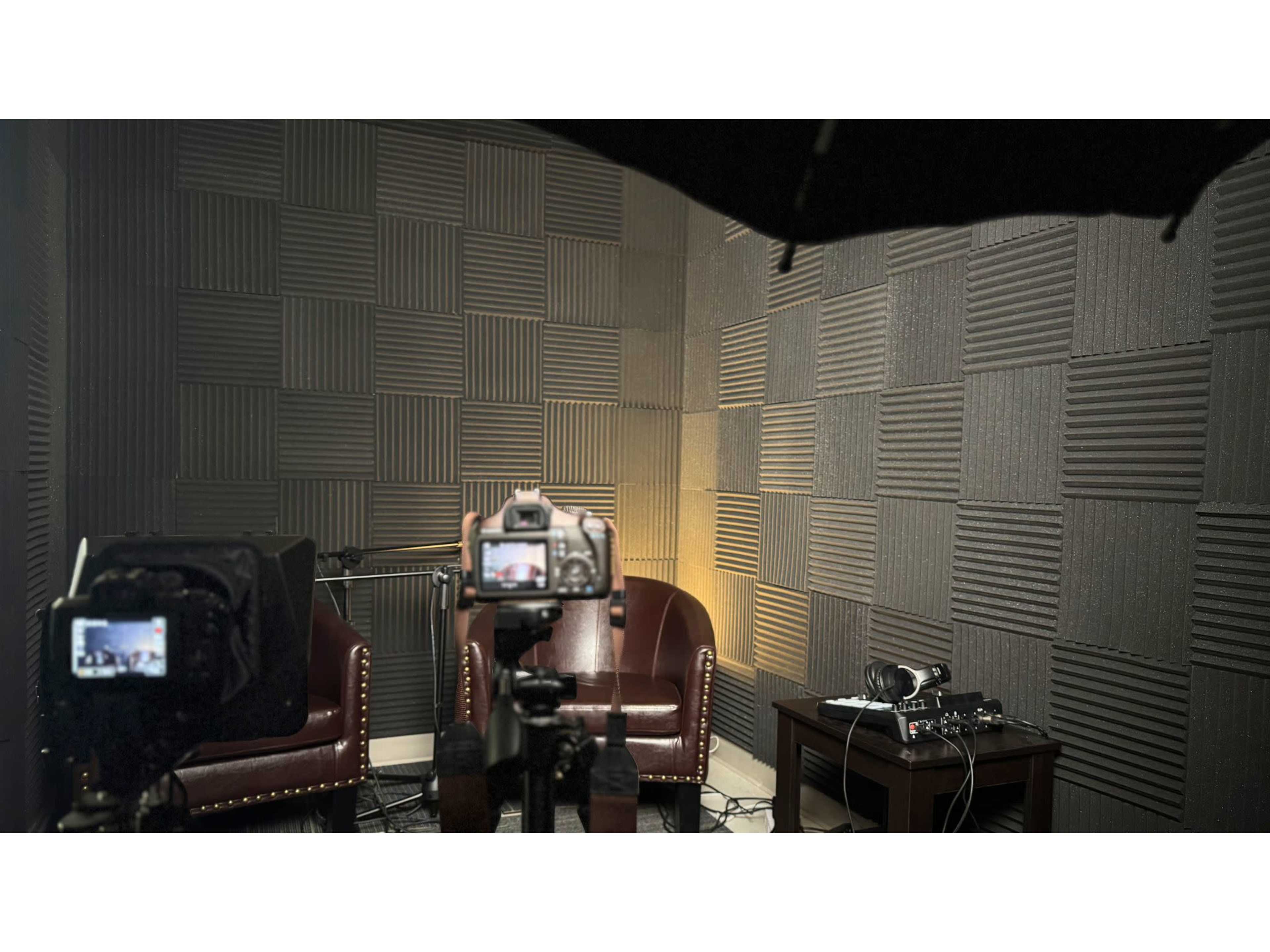 The image shows a soundproof recording studio featuring two leather chairs, cameras set up for filming, and audio equipment on a small table.