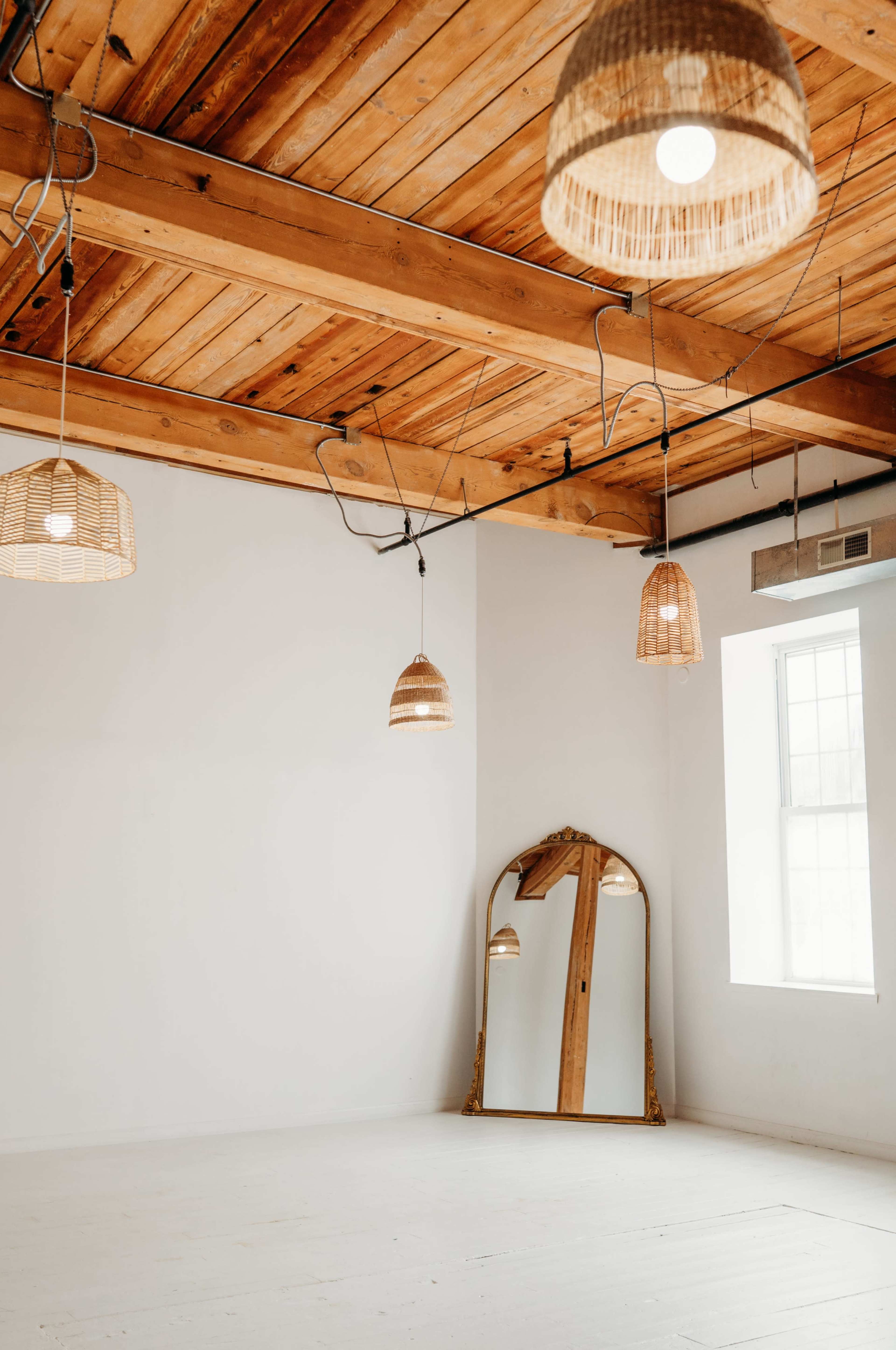 The image shows a spacious room with wooden beams on the ceiling, several pendant lights, and a large mirror leaning against a wall.