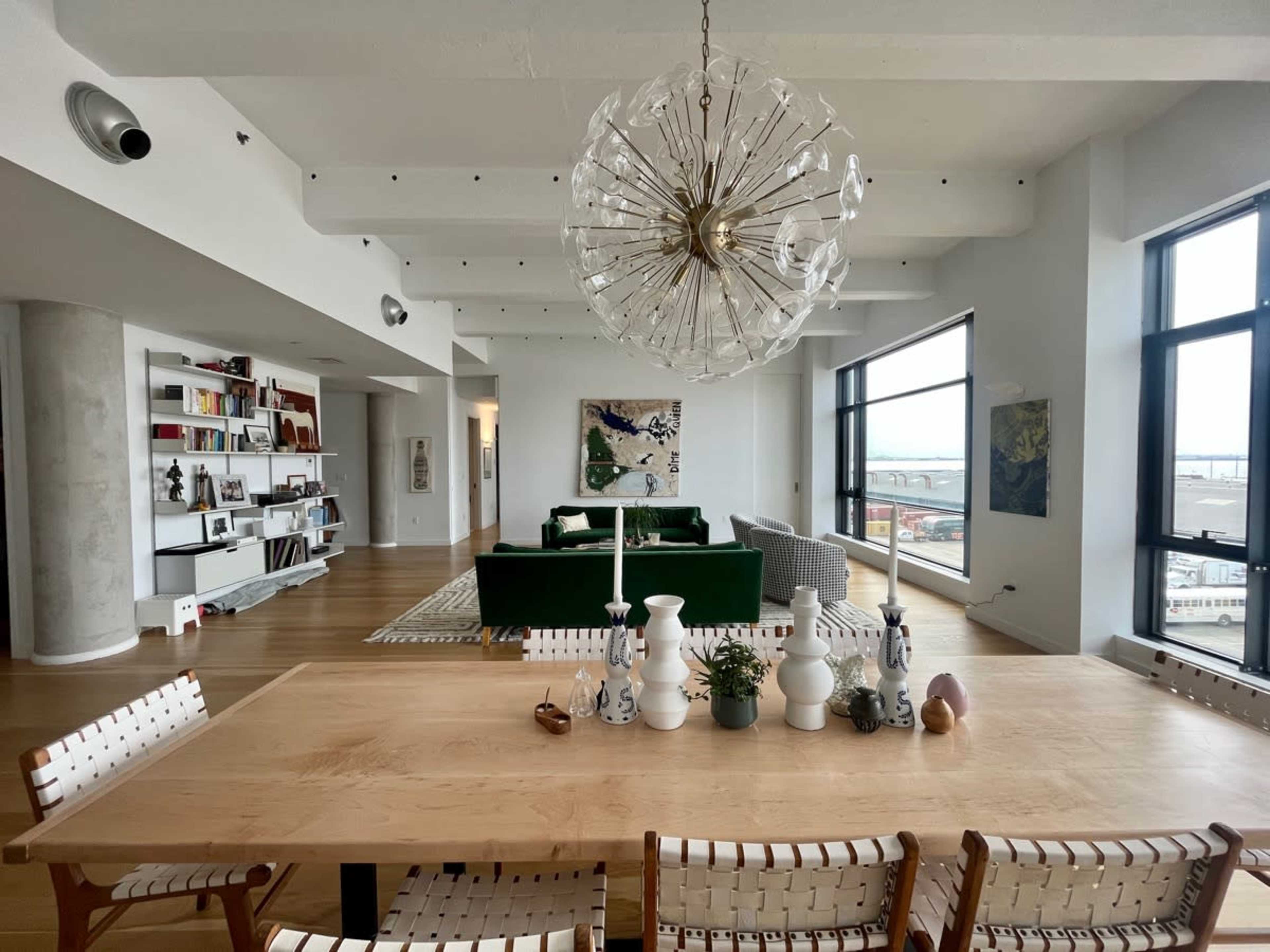 A modern living space featuring a wooden dining table with chairs, a green sofa in the background, large windows letting in natural light, and artistic decor on the walls.